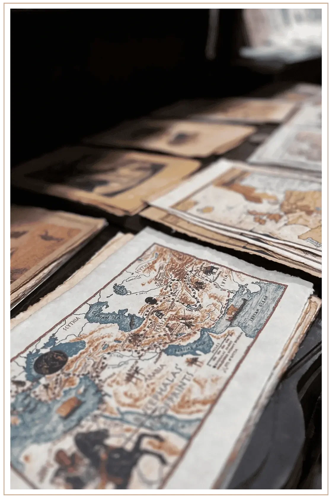A close-up, shallow-depth-of-field shot of antique maps on deckled-edge paper, showing historical illustrations of Scythia, Persia, and the Indian Ocean in muted earthy tones.