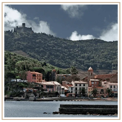 A moody, desaturated view of the historic Mediterranean town and fortress of Collioure, France, set into a terraced, vineyard-covered hill by the sea.