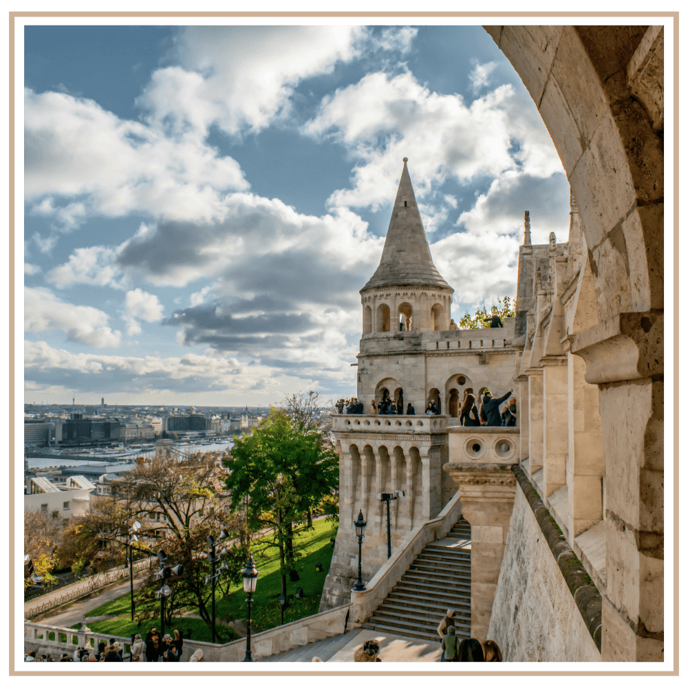 View from the stone arches of Fisherman’s Bastion in Budapest, featuring neo-Romanesque towers, a wide staircase, and a panoramic overlook of the city and Danube River under a blue sky with clouds.