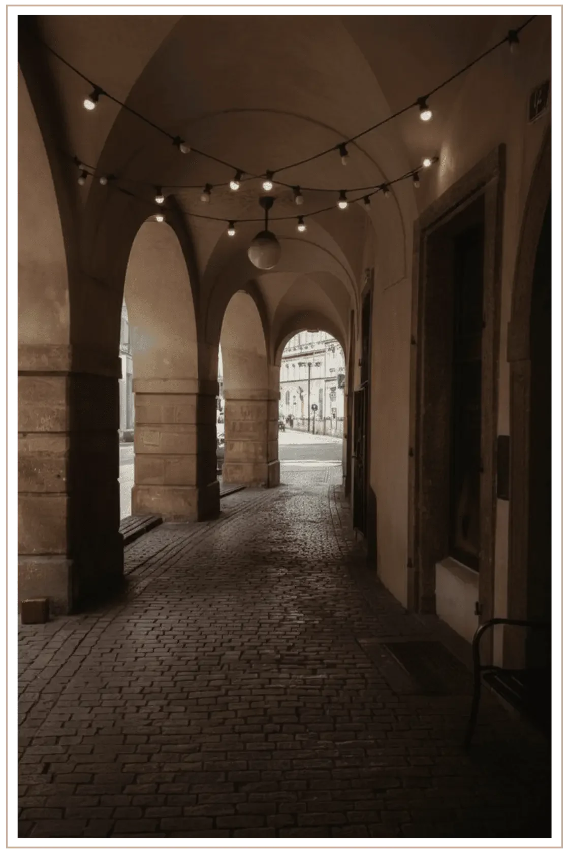 A moody, sepia-toned shot of a cobblestone arcade in Linz, Austria, featuring historic vaulted stone arches and hanging café string lights leading toward a sunlit town square in Linz, Austria.
