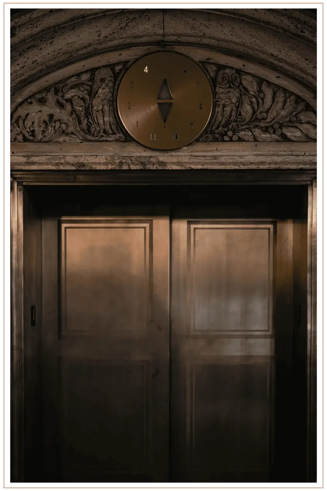 Close-up of vintage brass elevator doors in a luxury hotel, featuring an ornate stone archway with classical carvings of owls and a circular brass floor indicator dial.