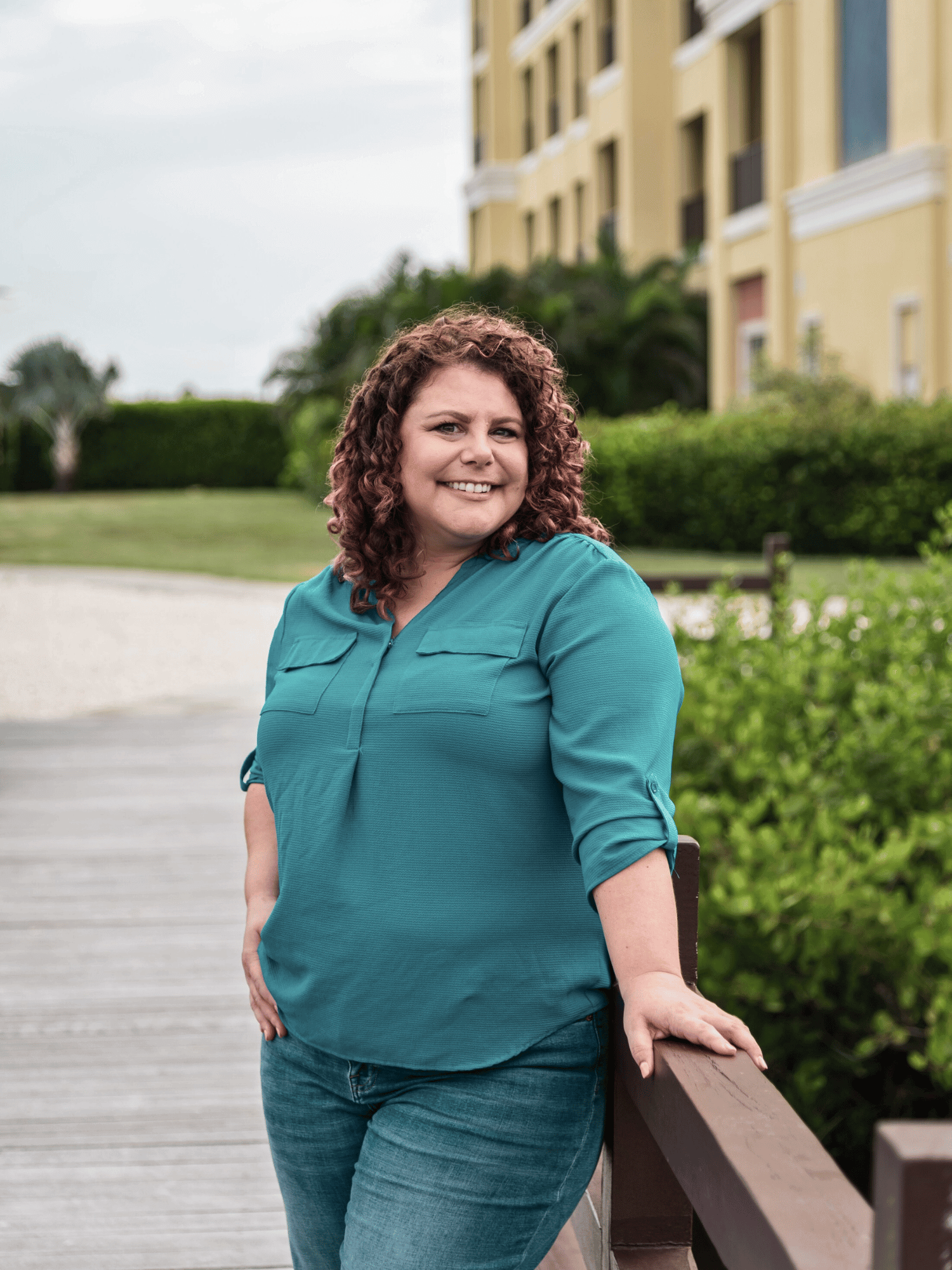 Denise Kubica wearing a teal blouse and jeans, smiling outdoors near a wooden railing, with a building and greenery in the background.