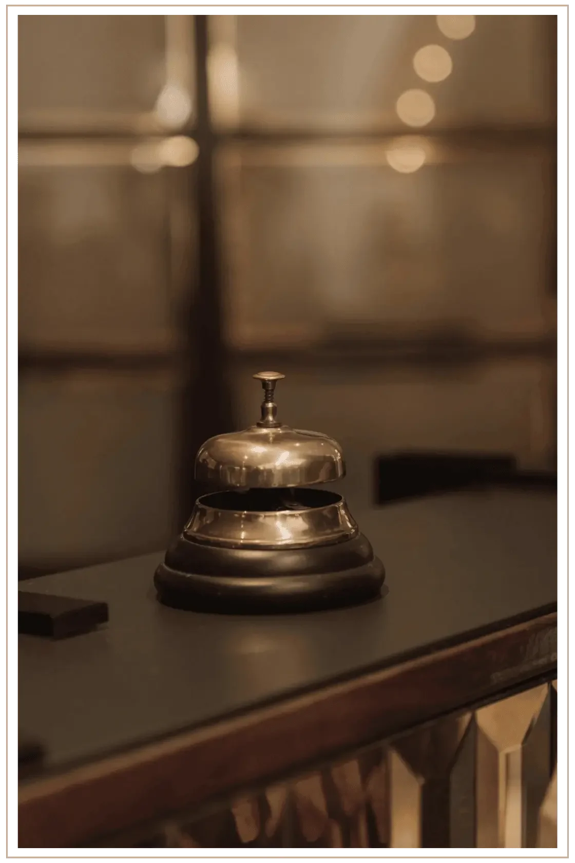 A close-up shot of a vintage brass concierge service bell resting on a dark, polished wood reception desk in a high-end hotel, with a blurred, warm-toned interior in the background.