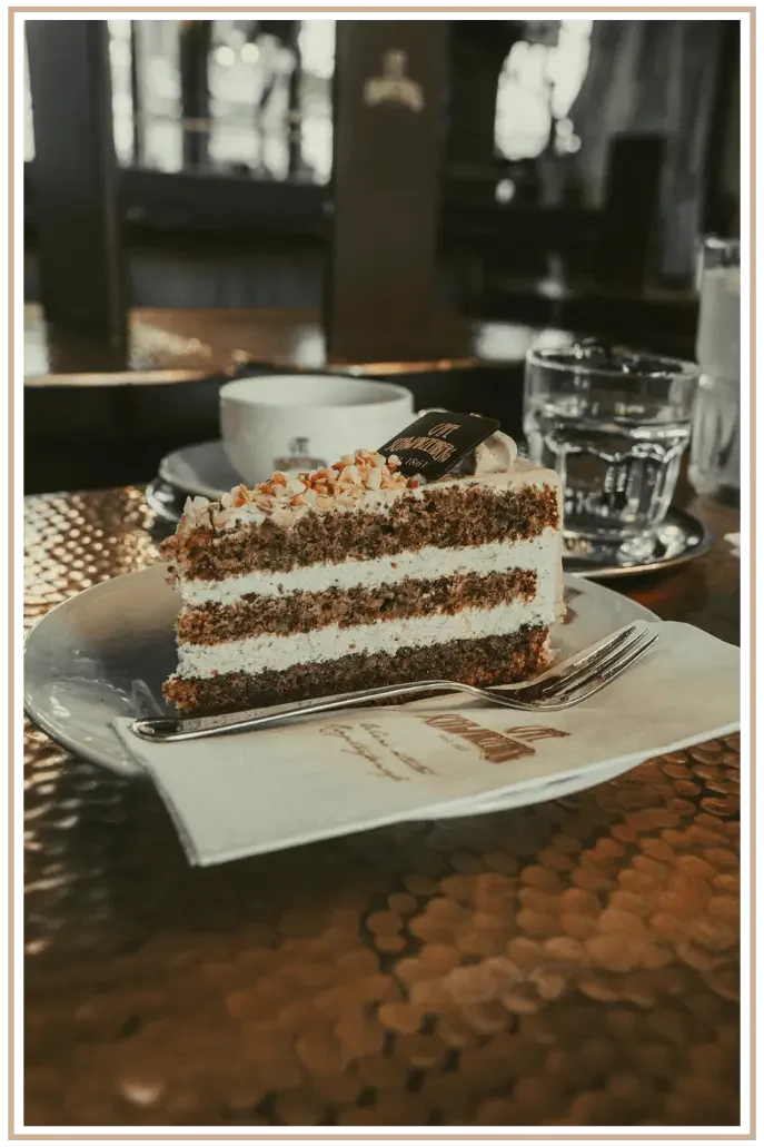 A photograph focused on a slice of a delicious layered cake in a Vienna cafe setting. A water glass and coffee cup are on the table, slightly out of focus.