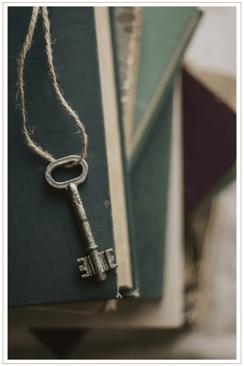 Close-up of an antique iron skeleton key hanging by a rustic twine loop against a stack of vintage cloth-bound books in shades of deep teal and forest green.