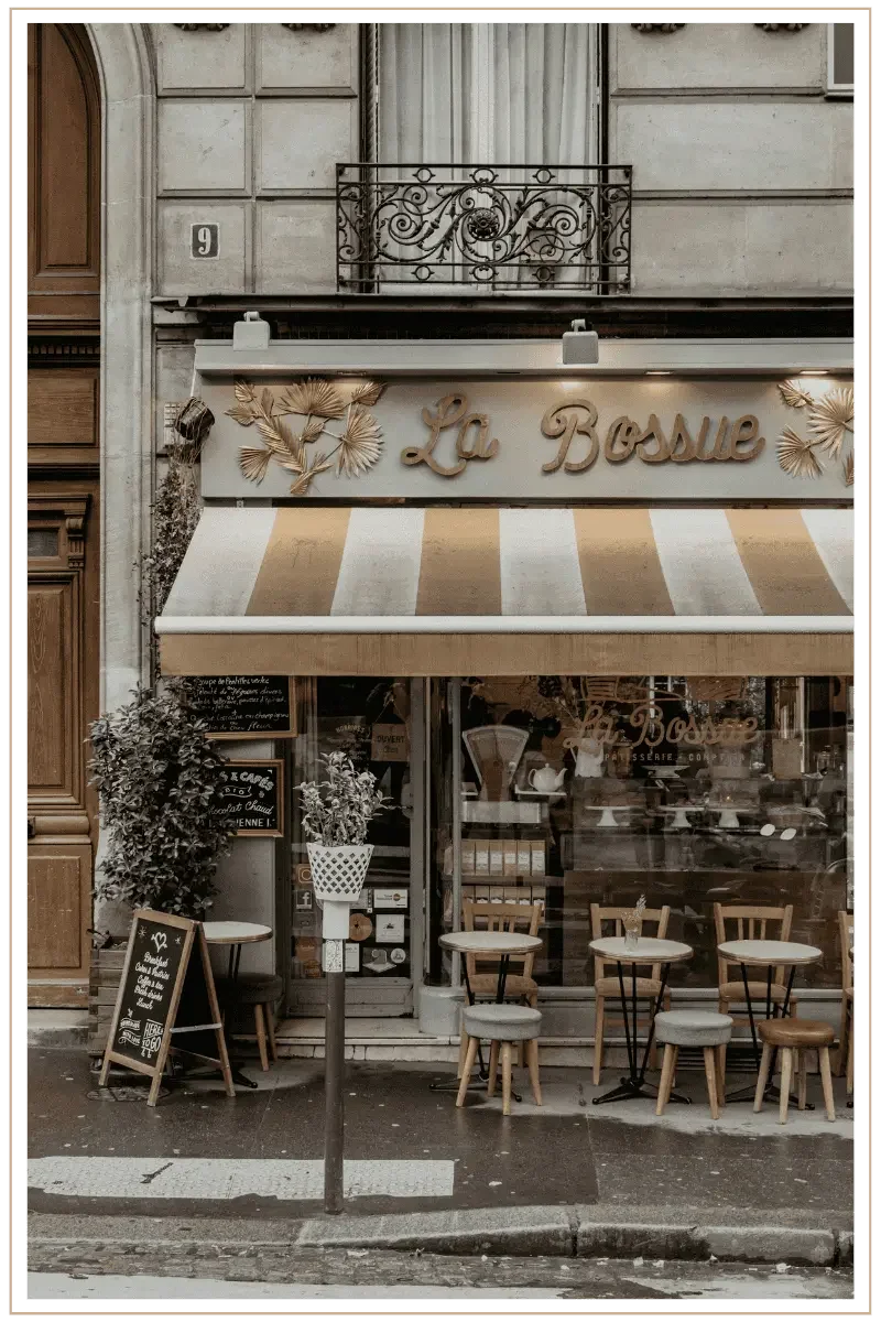 The outside of La Bossue cafe in Paris, France. Small tables with chairs are placed out front, with a small chalk sign placed next to the door. A decorative iron balister is above.