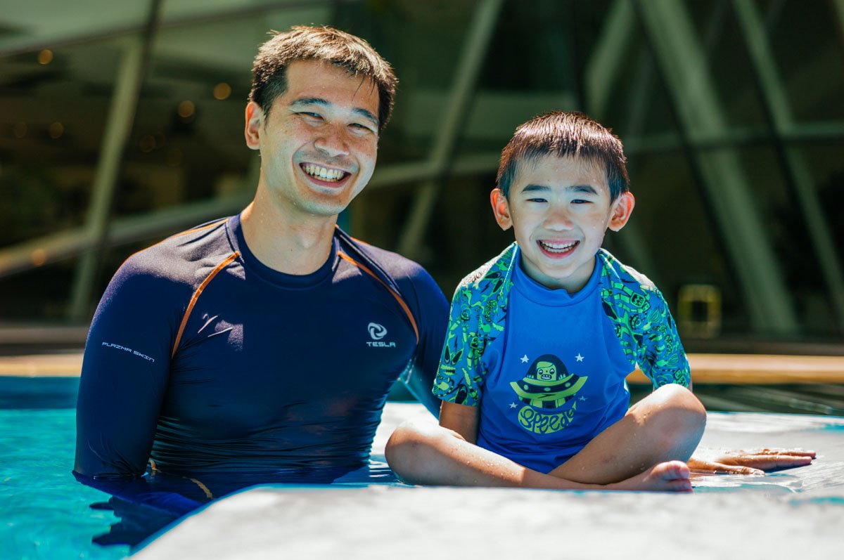 A man and a young boy smiling and sitting in a pool outdoors, with modern glass building in the background.