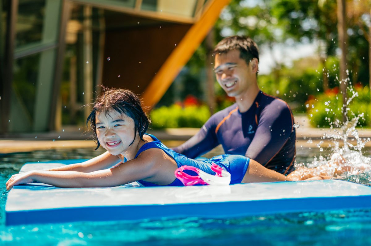 A young girl learning to swim with her instructor in a swimming pool on a sunny day. The girl is smiling and holding onto a pool float while the instructor supports her from behind.
