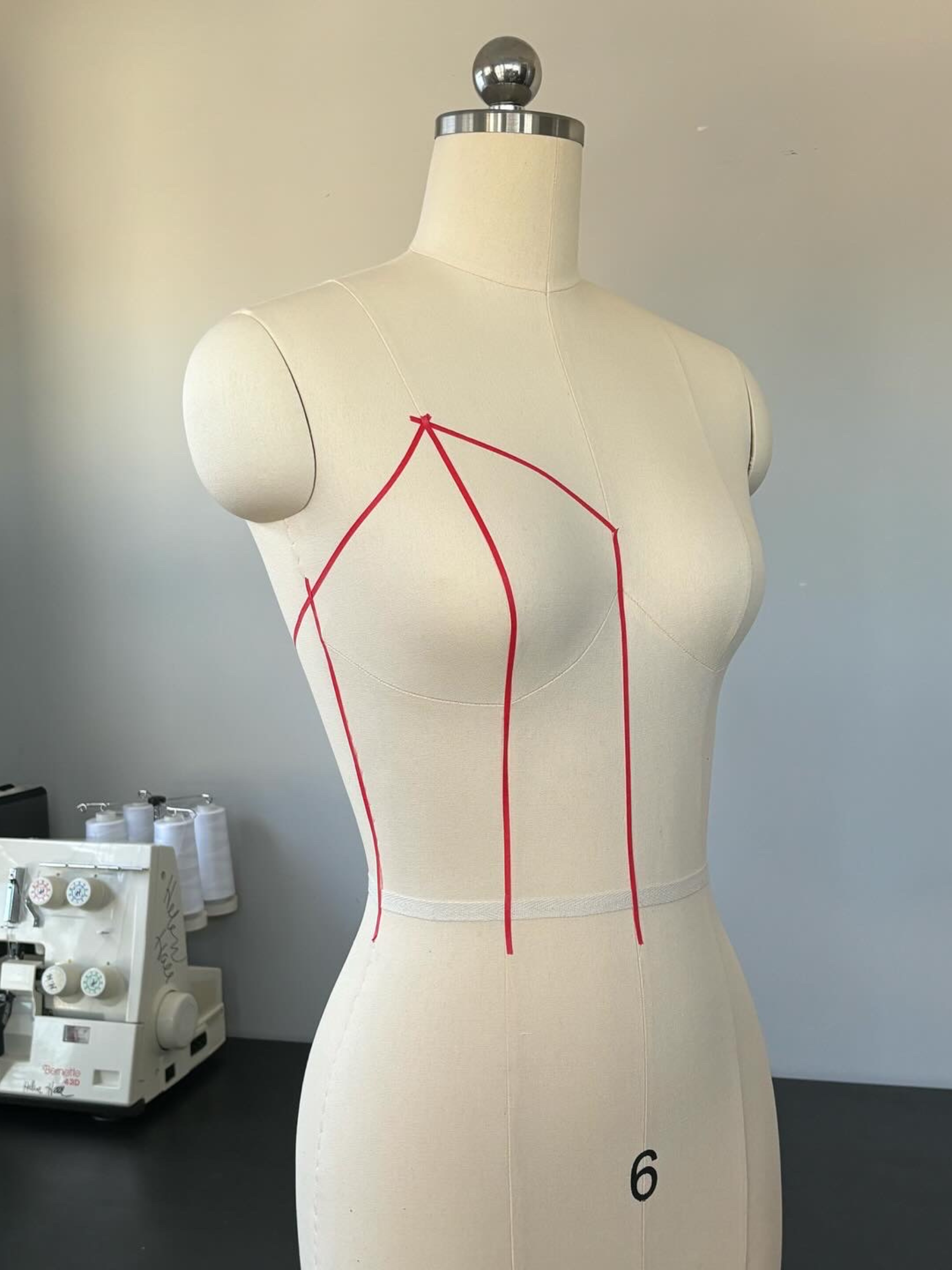 Dress form with red chalk markings indicating sewing lines, numbered '6' at the waist, in a sewing studio with sewing machine and spools of thread in the background.