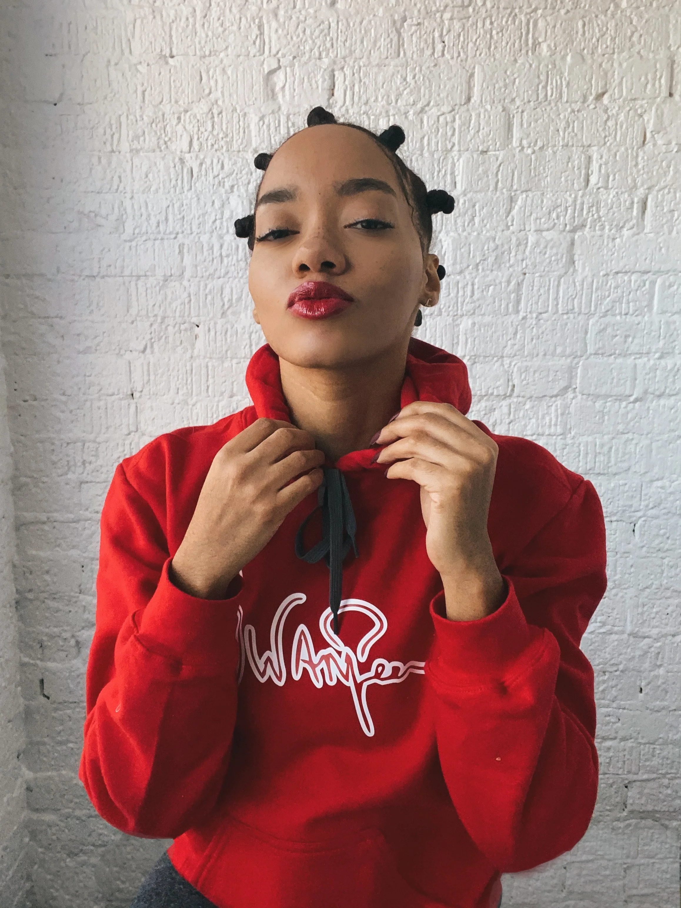 Young woman with bantu knots hairstyle wearing a red hoodie with white graphic text, standing against a white brick wall, looking confidently at the camera.