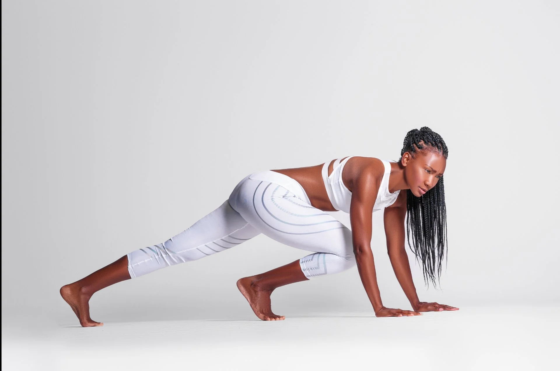 A woman in white athletic clothing practicing yoga on a plain white background, in a plank pose with one knee bent, one arm extended, and her head lowered.