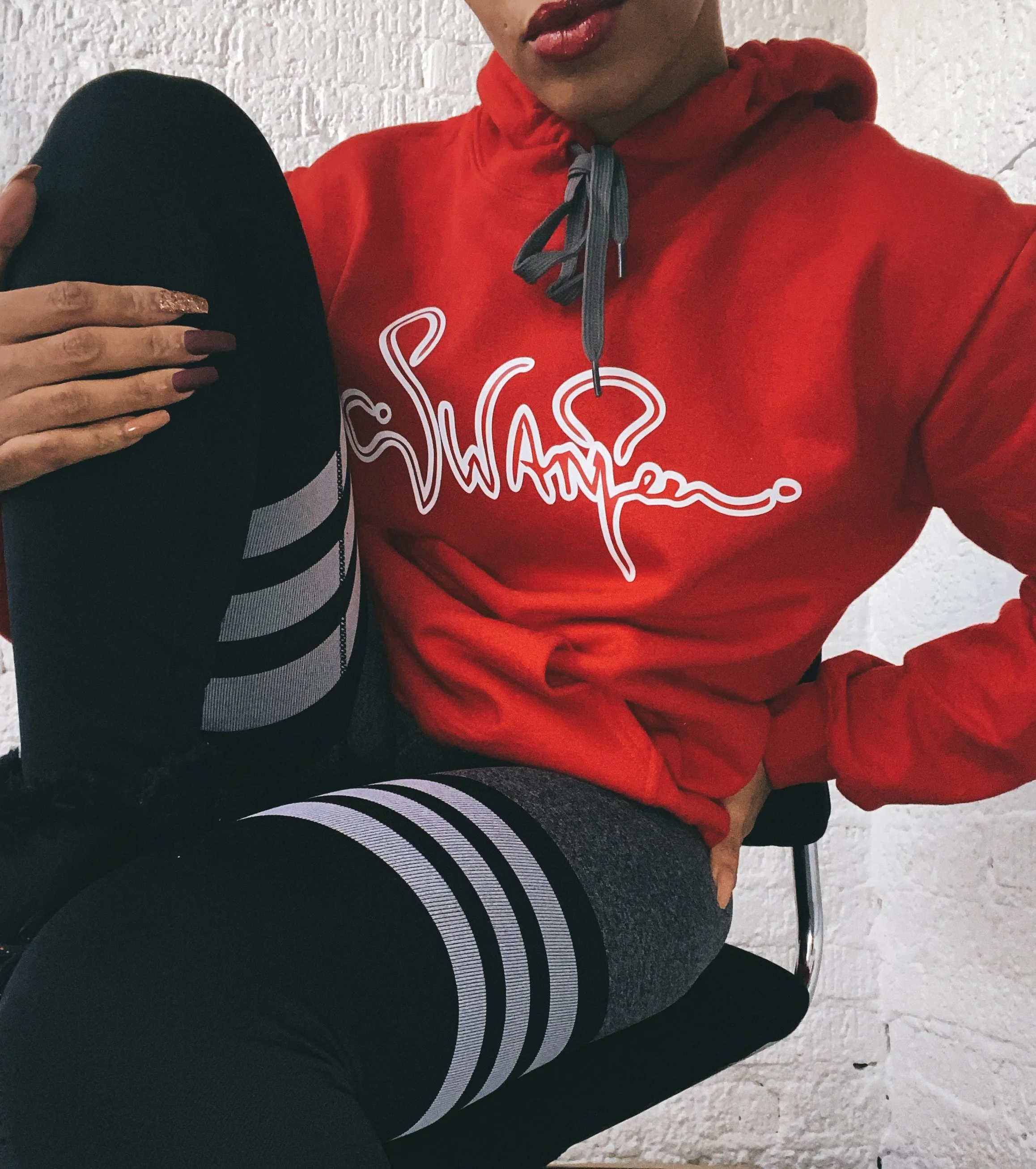 Person wearing a red hoodie with white lettering and black shorts with gray stripes, sitting on a chair against a white brick wall.