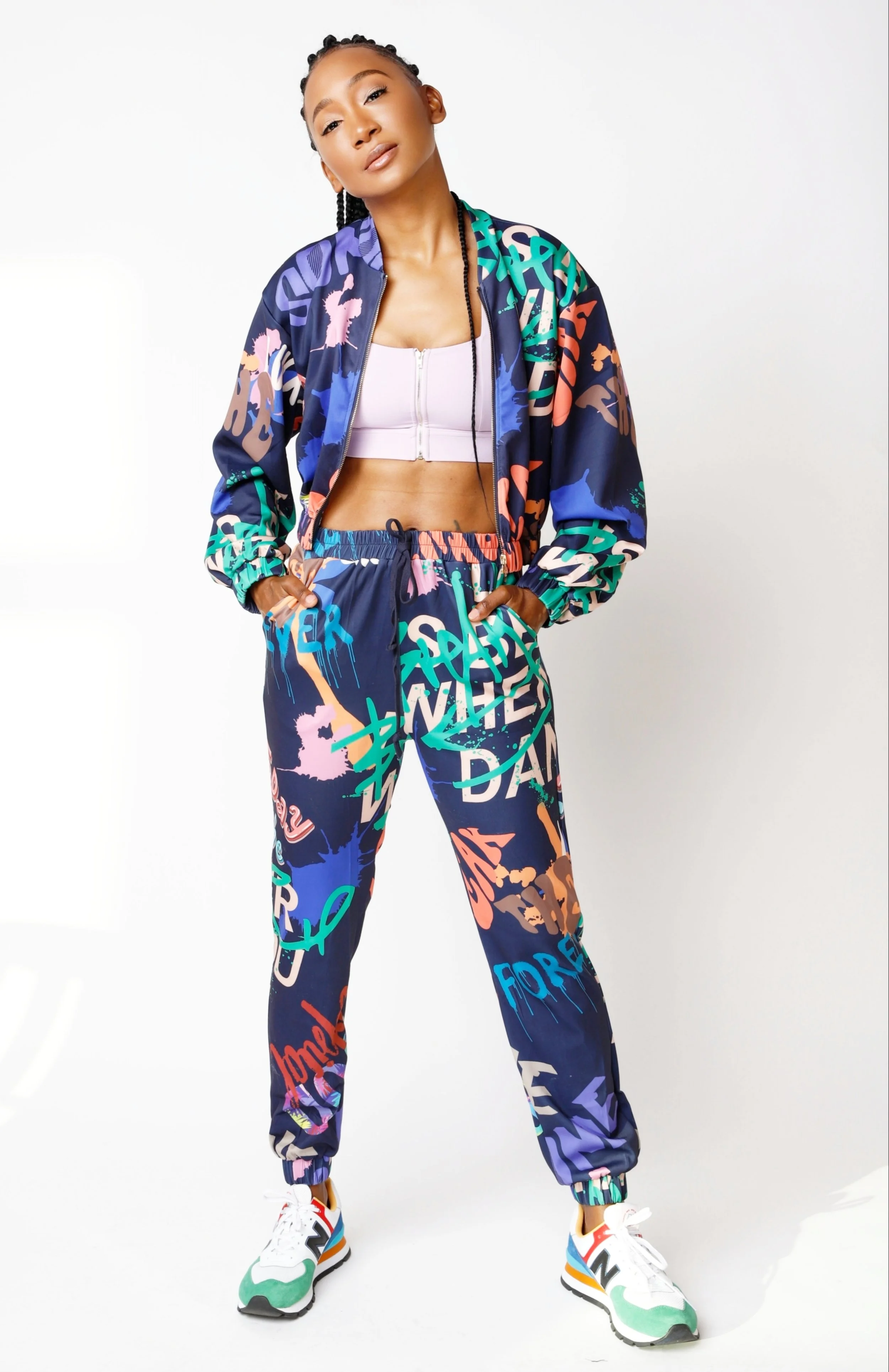 A woman wearing a colorful graffiti-patterned matching jacket and pants, a white crop top, and sneakers, standing against a plain white background.