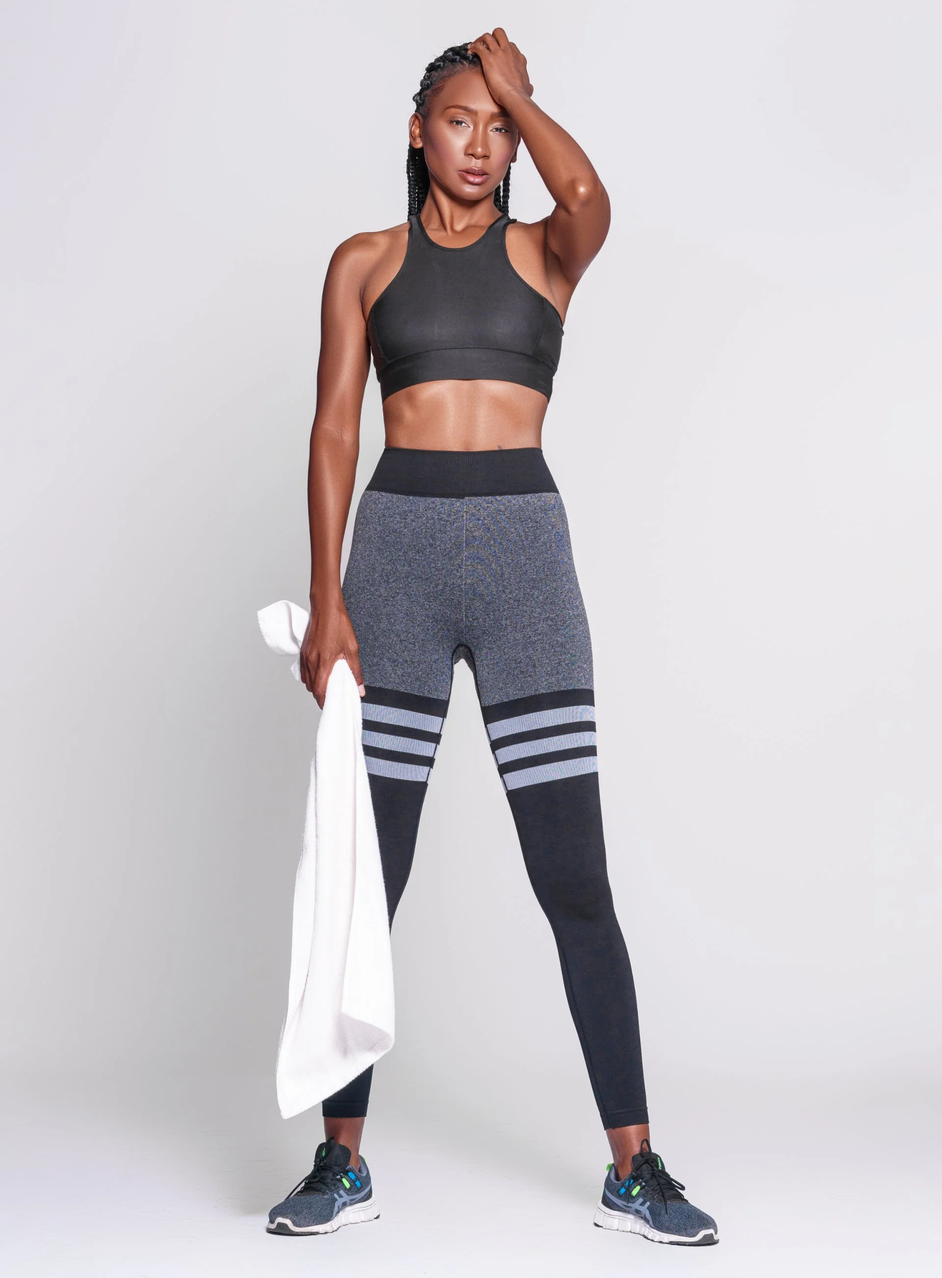 A woman in athletic wear standing against a plain light gray background, holding a white towel in her right hand, with her left hand resting on her head, and looking at the camera.