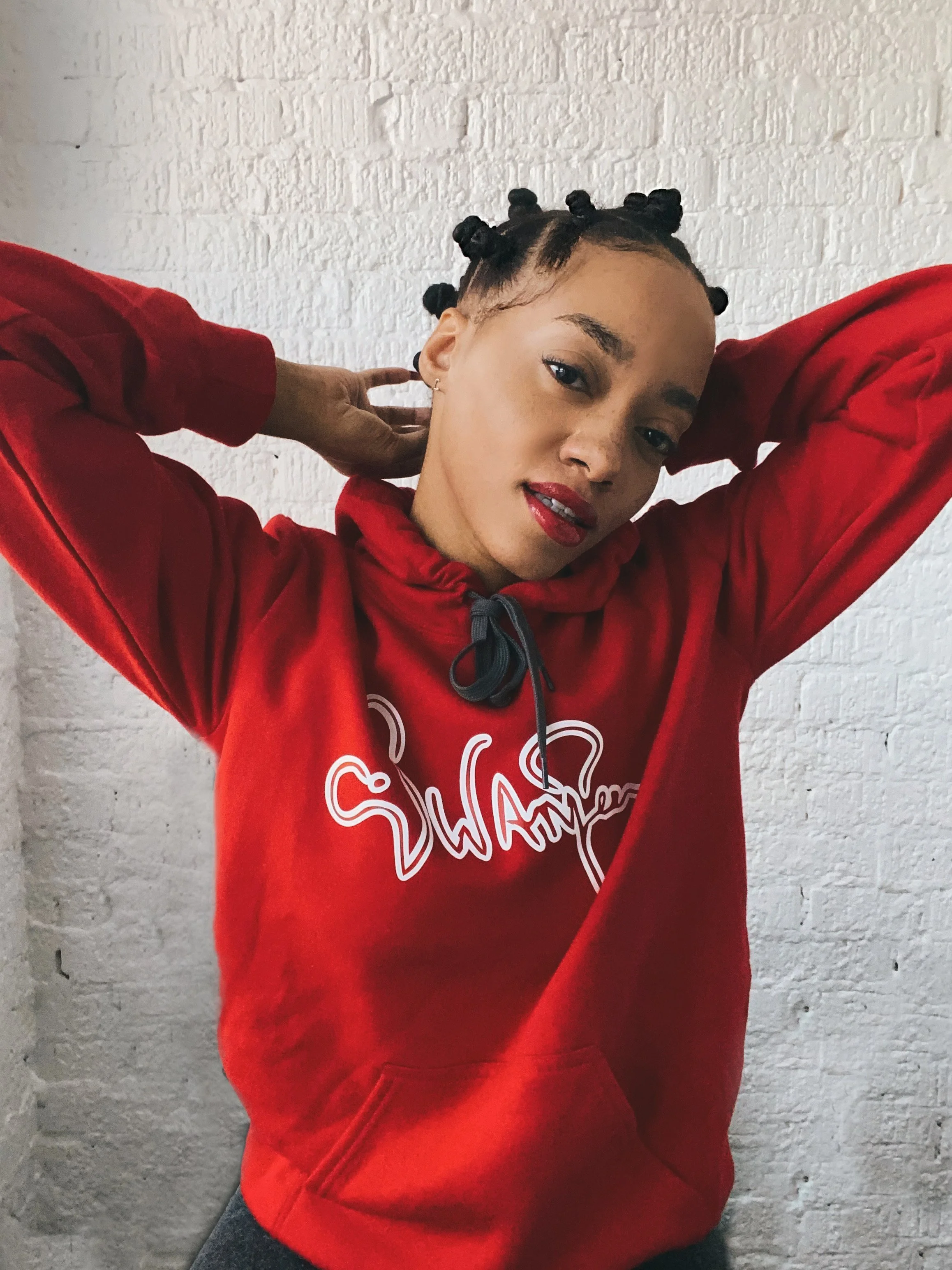 A young woman with her hair styled in Bantu knots, wearing a red hoodie with white writing, posing with her hands behind her head against a white brick wall.