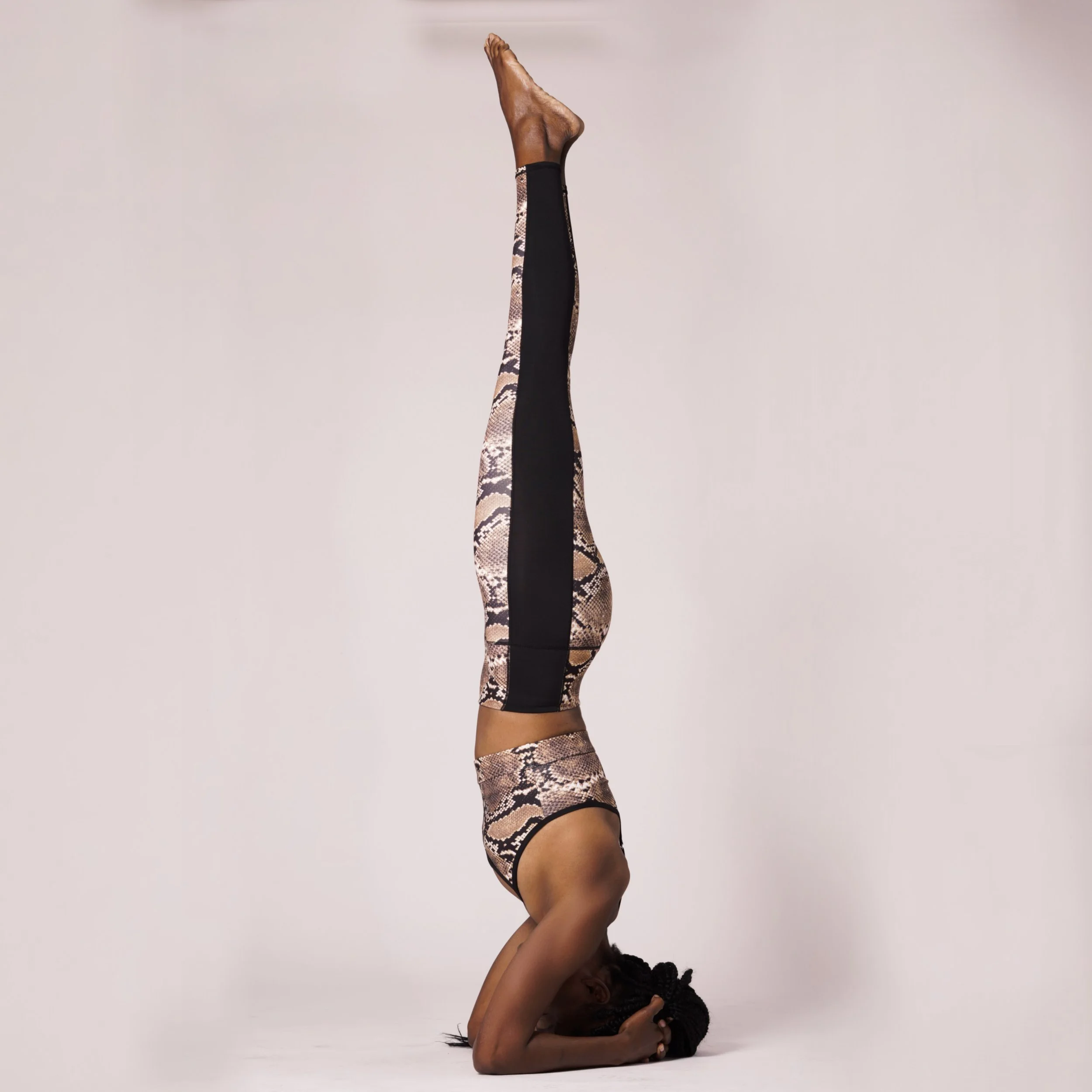 A woman performing a headstand yoga pose against a plain white background, wearing a snakeskin pattern workout outfit and brown ankle boots.