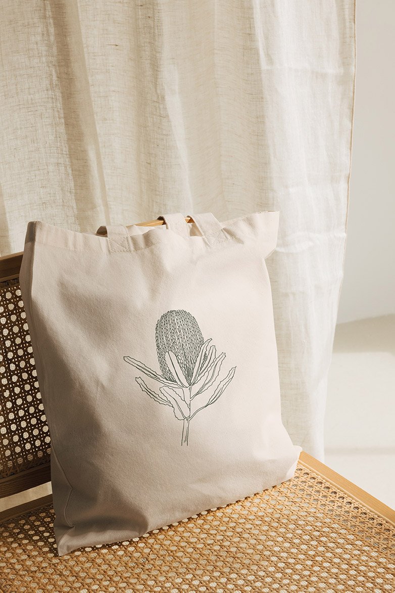 A beige tote bag with a line drawing of a banksia on it, resting on a woven chair near light-coloured curtains.