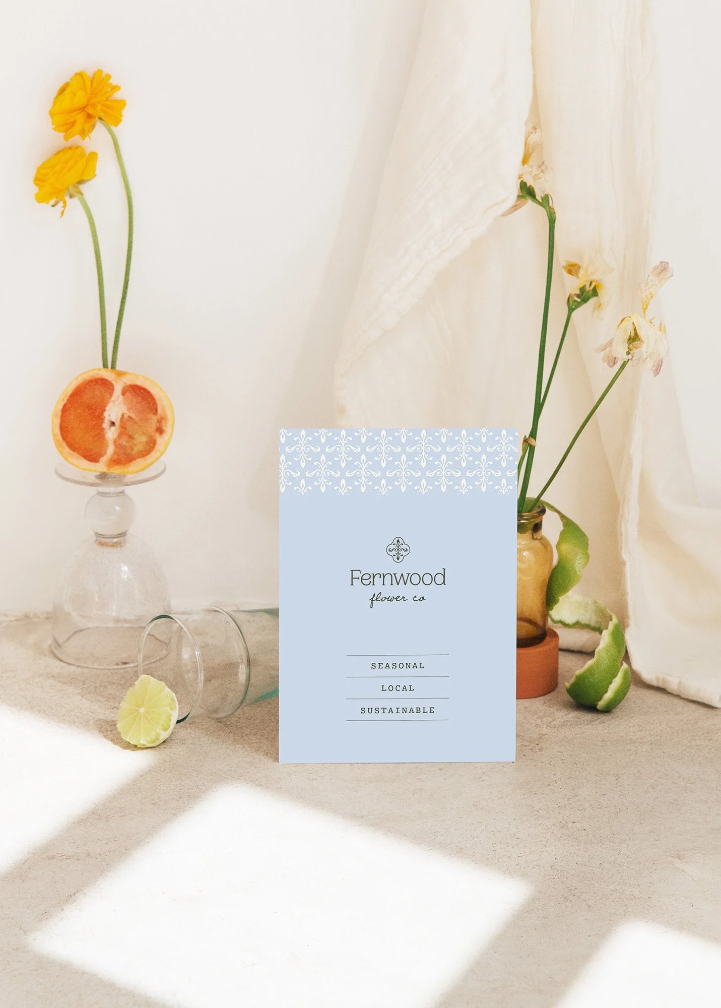 A bouquet of dried flowers and citrus slices arranged on a beige surface, with a blue Fernwood flower co brochure in the foreground. The background includes a white curtain and a potted plant.