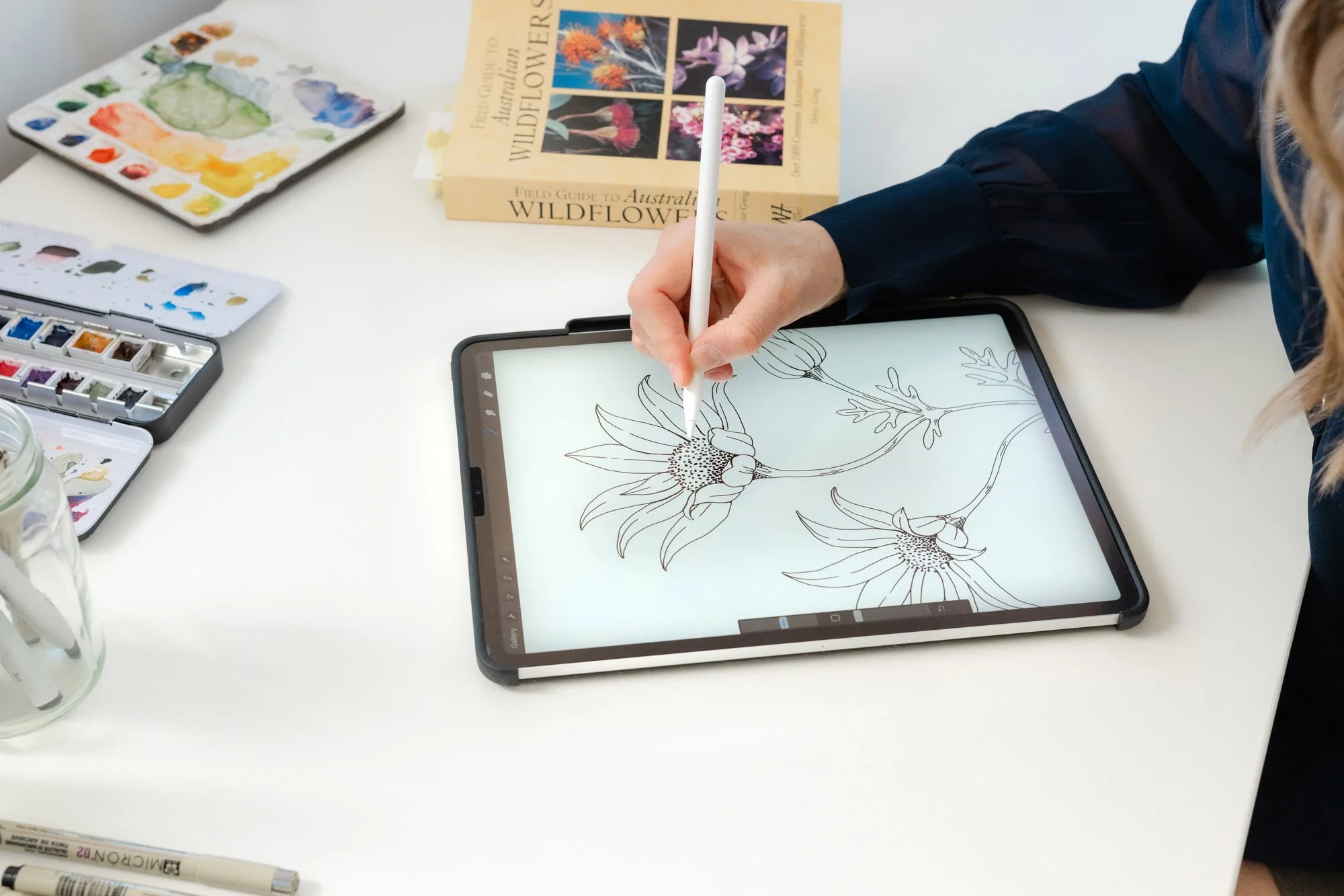 A person is drawing a detailed floral illustration of flowers on an iPad. The workspace has watercolor paints, brushes, and a botanical book titled 'Field Guide to Australian Wildflowers' on the table.