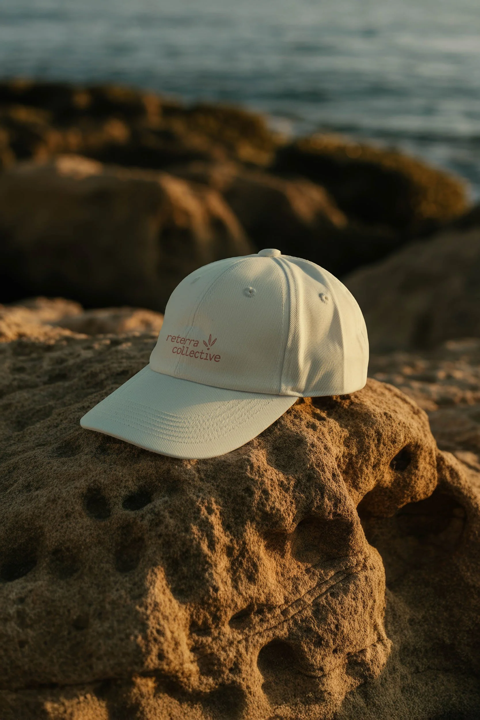 A white baseball cap with the words "reterra collective" embroidered on the front, resting on a large textured rock on a beach near the ocean, with rocks and water in the background during sunset.