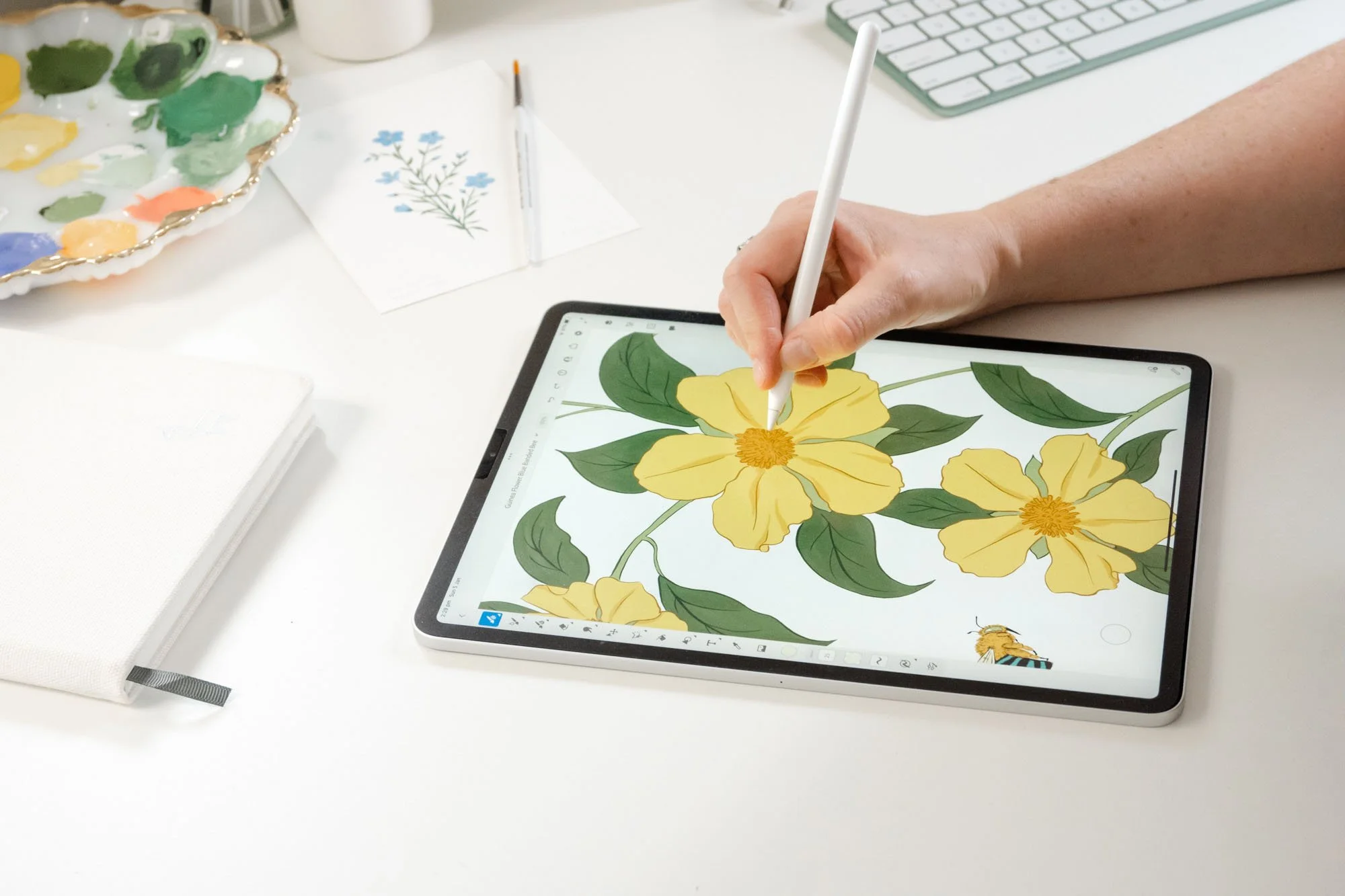 Person drawing yellow flowers with green leaves on a digital tablet using a stylus, surrounded by art supplies and a keyboard.