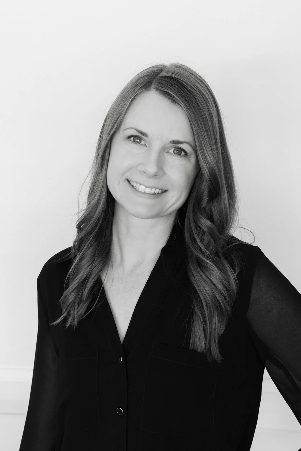 Black and white portrait of a woman with long wavy hair, smiling, wearing a black button-up shirt, standing against a plain white background.