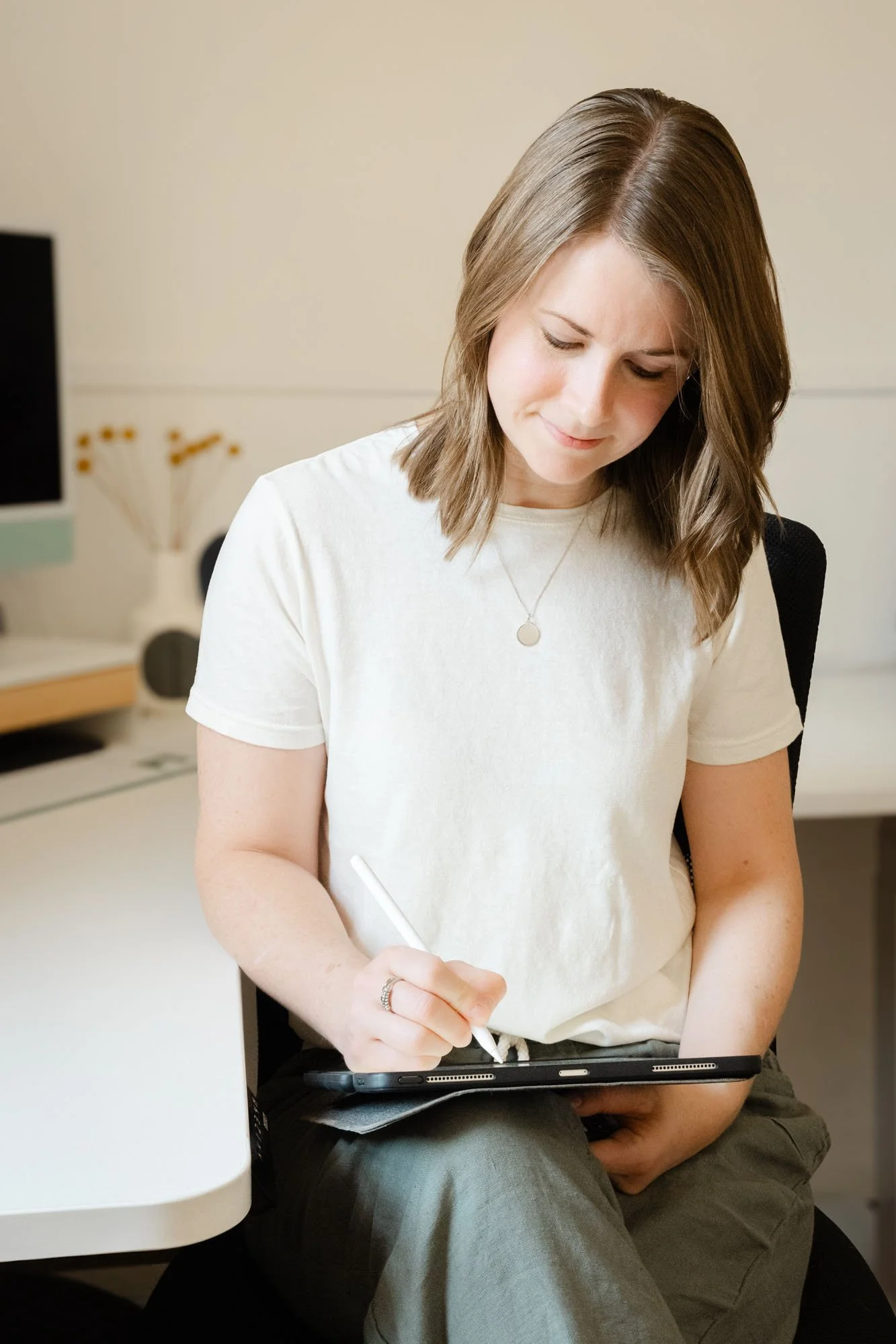 Woman sitting at a desk, using a stylus on a tablet, with a neutral expression.