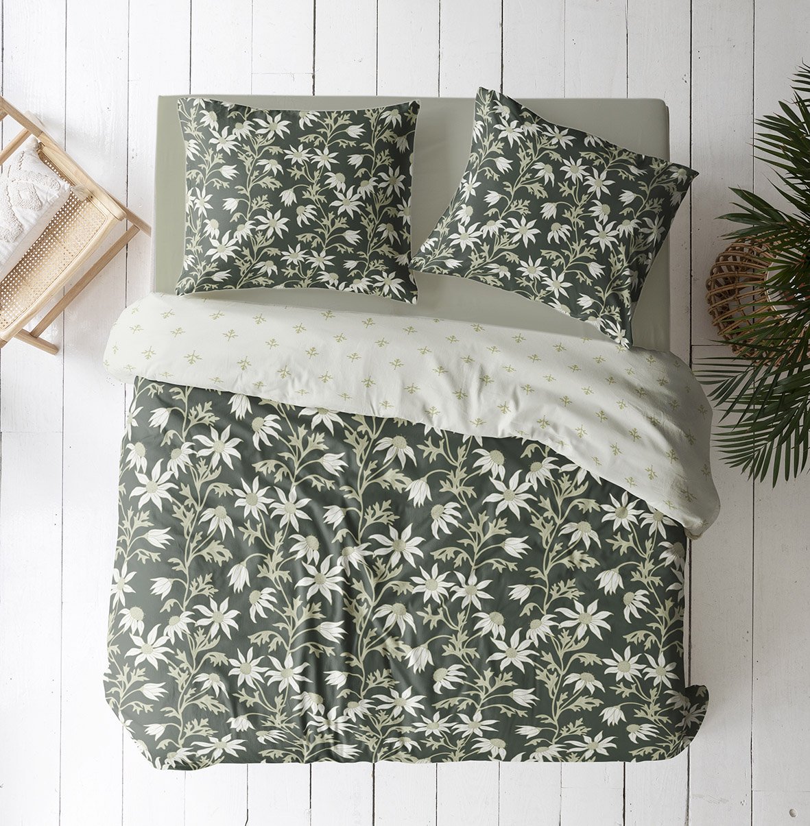 A bed with black and white daisy-patterned bedding, green pillows with white floral print, a white sheet with small green starburst design, a small wooden chair with a white cushion to the side, and a leafy green plant on the right.