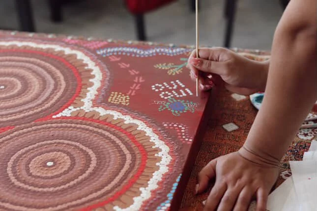 Close-up of a person creating a dot painting on a red canvas, using a small stick to apply white and other colored dots in circular patterns.