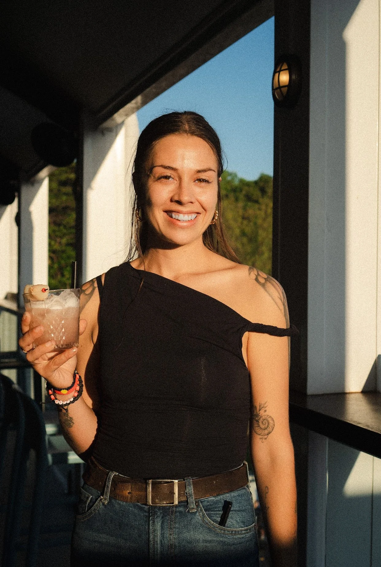 A woman smiling and holding a cocktail with a cherry garnish in her right hand, standing near a window with sunlight on her face and a blue sky in the background.