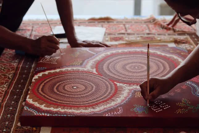 Two people creating a dot painting on a square canvas, featuring concentric circles and colorful patterns, on a woven rug surface.