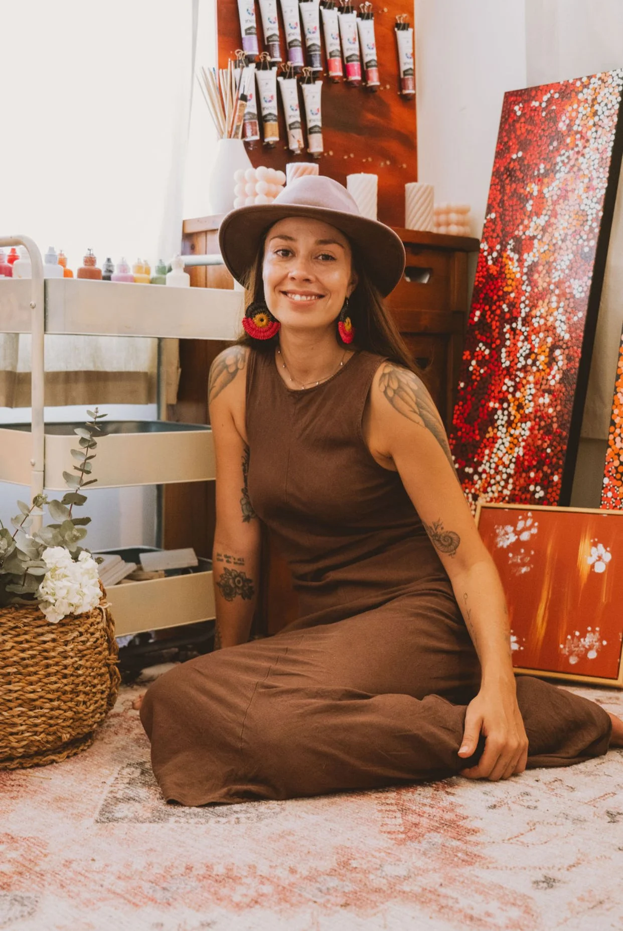 A woman smiling and sitting on the floor in front of a display of colorful art supplies and paintings, wearing a brown dress, a lavender wide-brimmed hat, and colorful earrings.