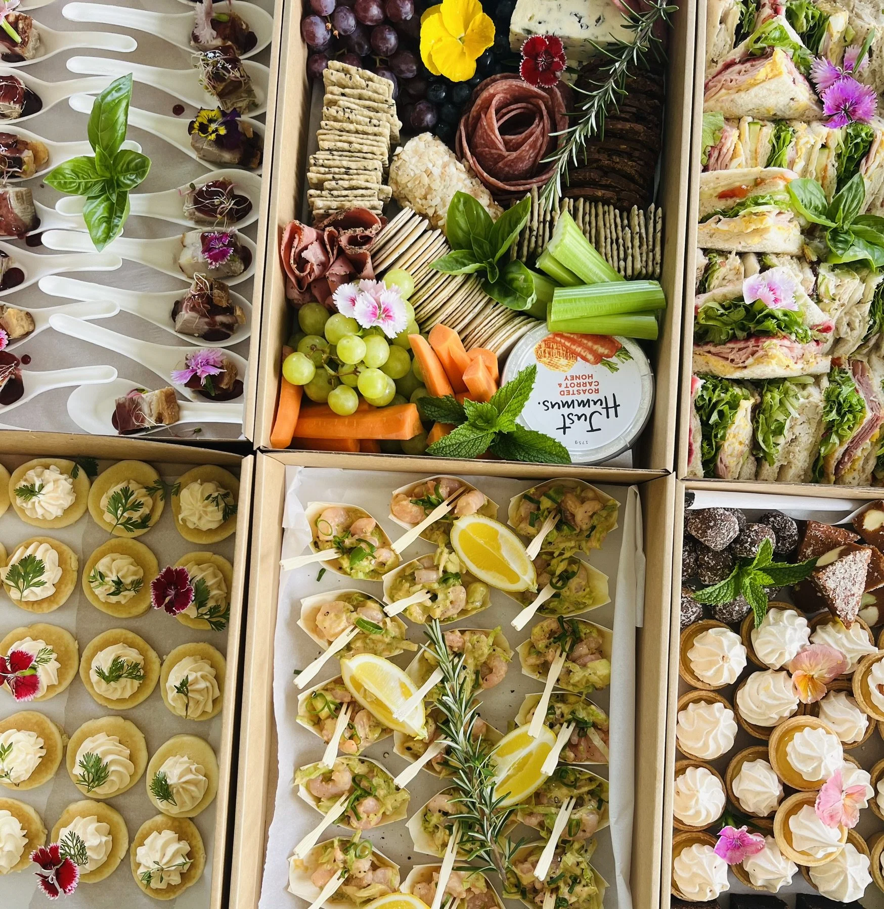An assortment of appetizers and desserts arranged in wooden boxes, including mini tartlets with herbs, shrimp bites with lemon, assorted sandwiches, cheese and crackers with grapes, vegetables with dip, and small chocolates topped with pink flowers a