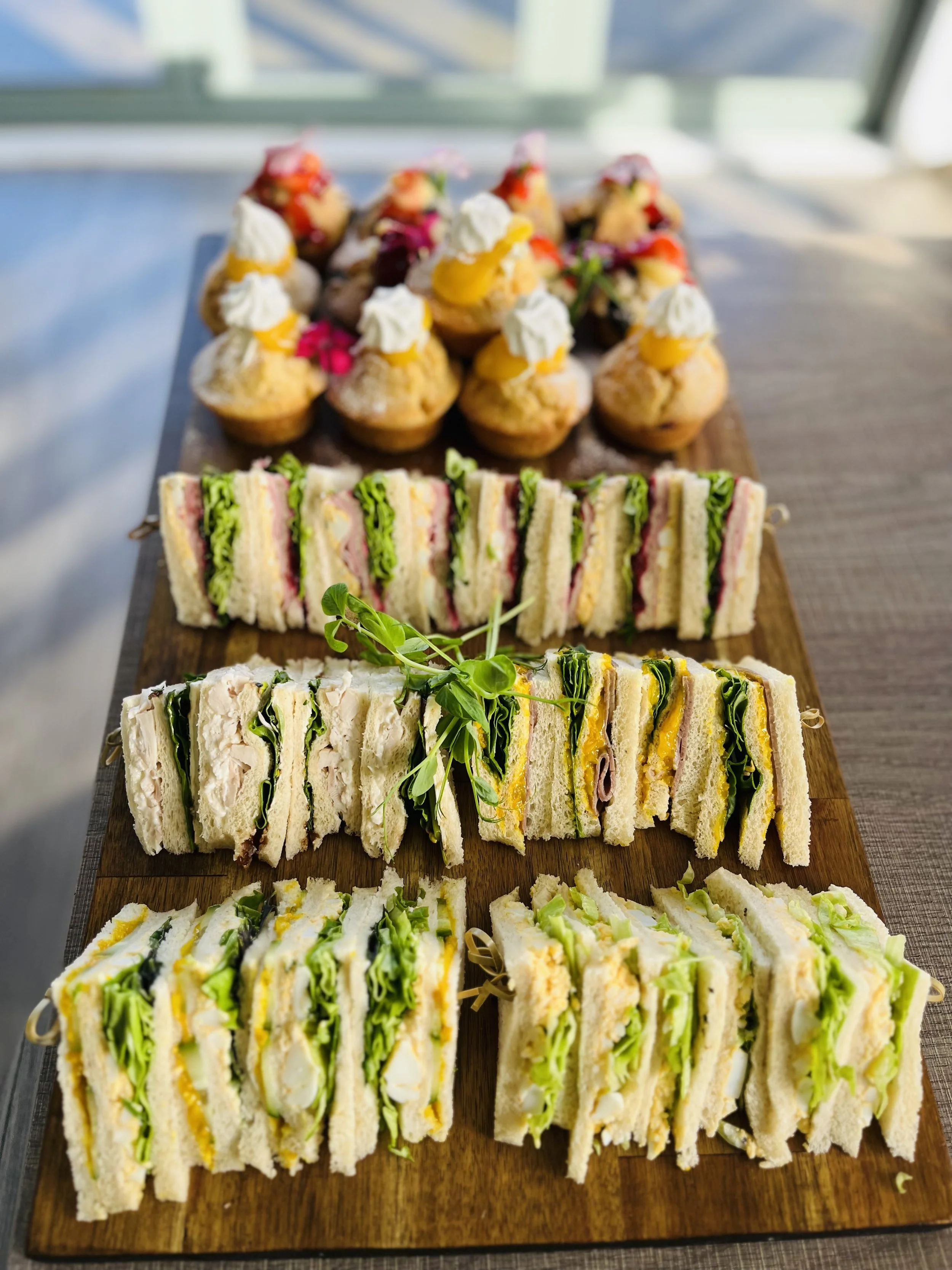 Assorted finger sandwiches and mini cupcakes on a wooden serving board.