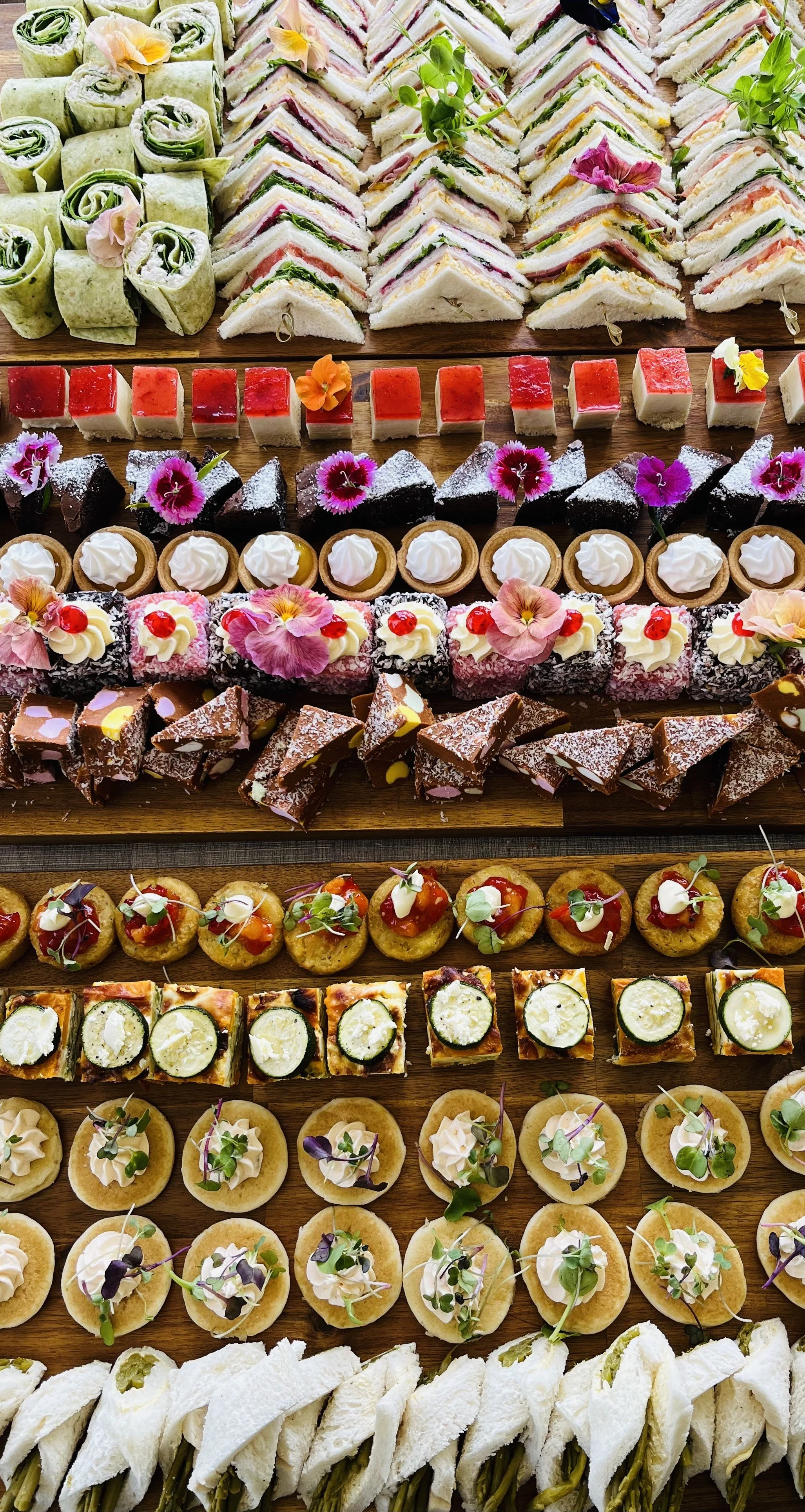 A colorful display of various finger sandwiches, mini cakes, sweets, and desserts arranged on wooden trays, decorated with edible flowers and microgreens.