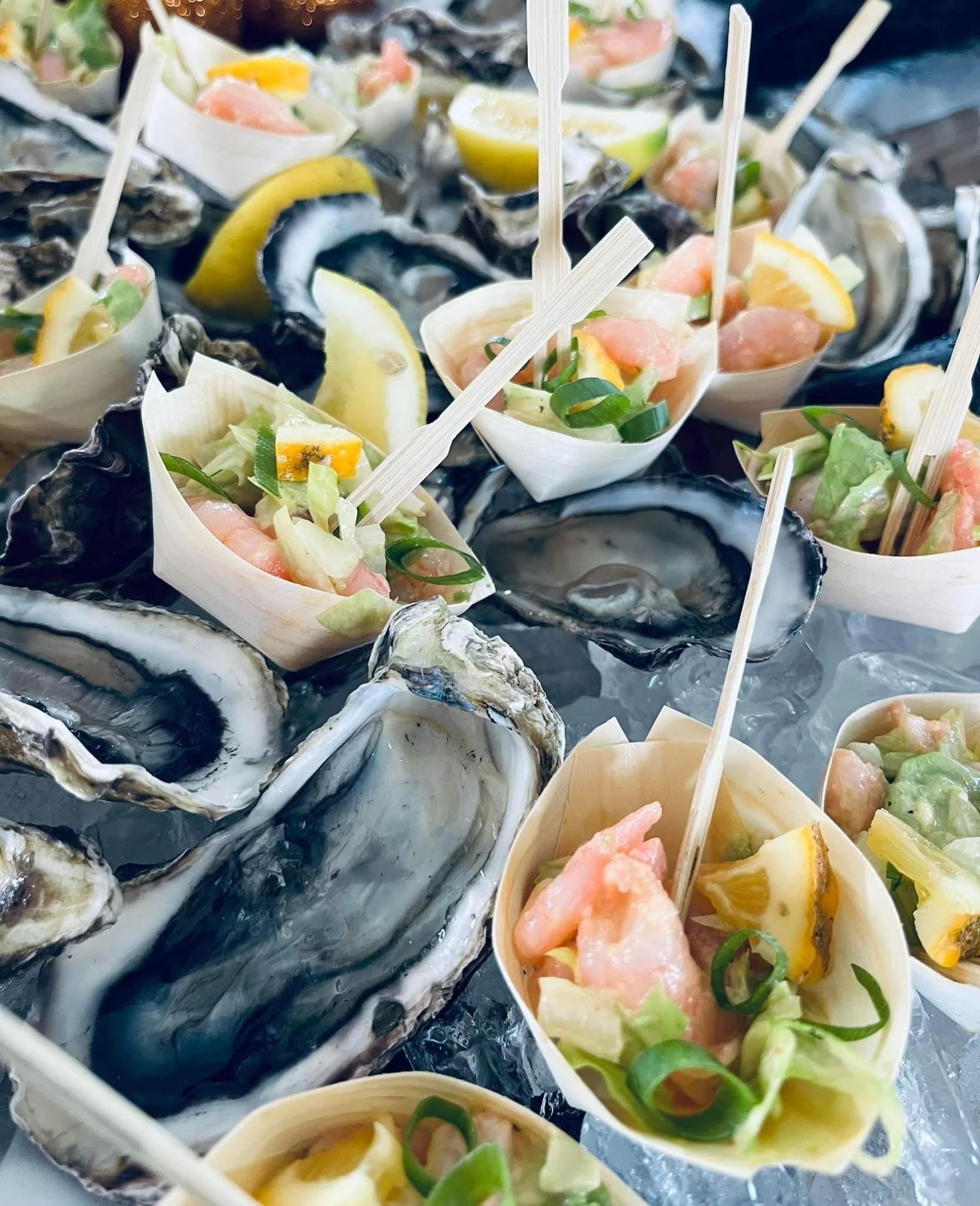 A display of fresh oysters served on ice with small cups of seafood salad, lemon wedges, and green onions.