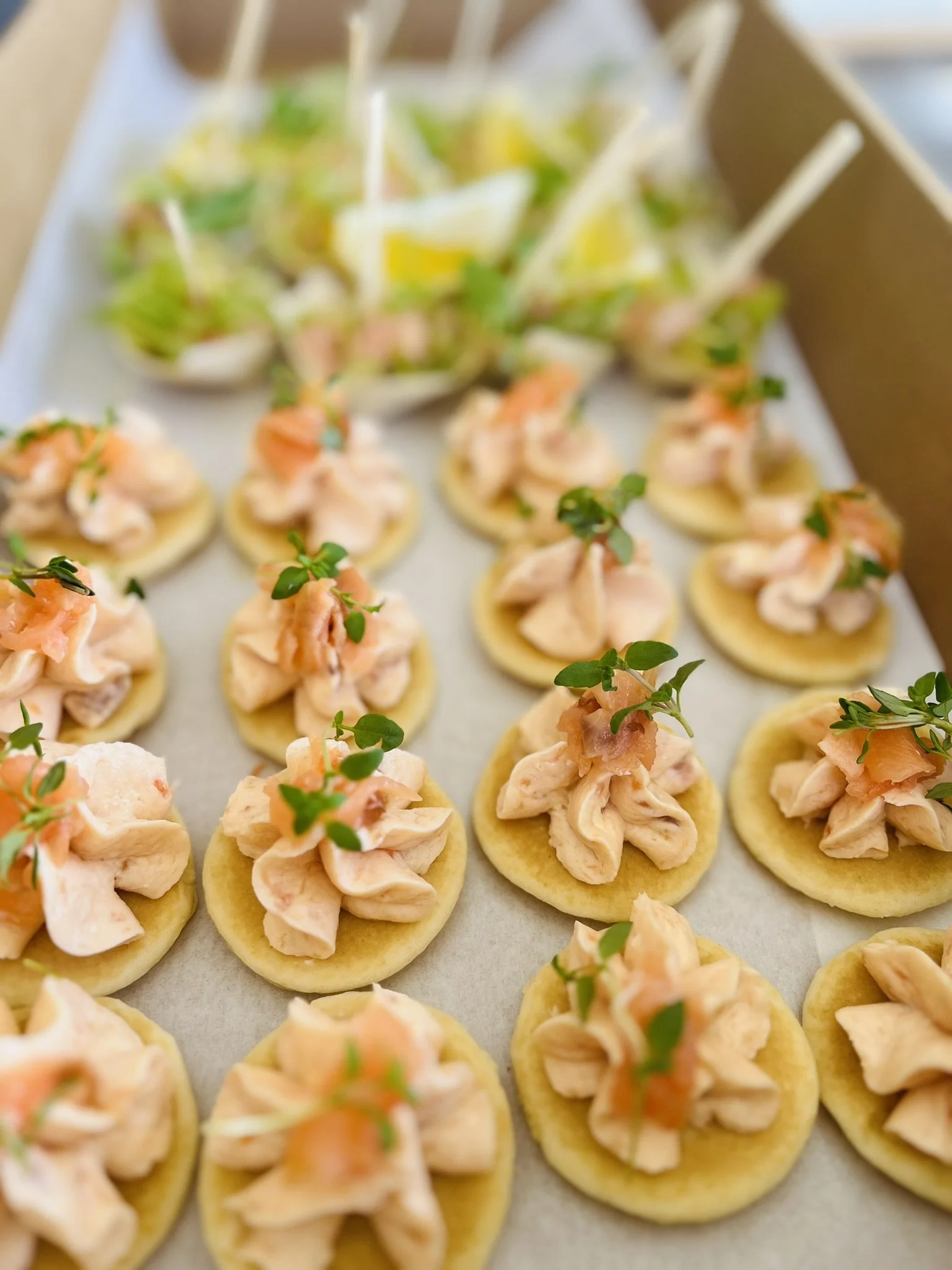 Small appetizer bites with pinkish chicken salad on round crackers, garnished with green herbs, arranged on a tray with a tilting container of small cups of salad in the background.