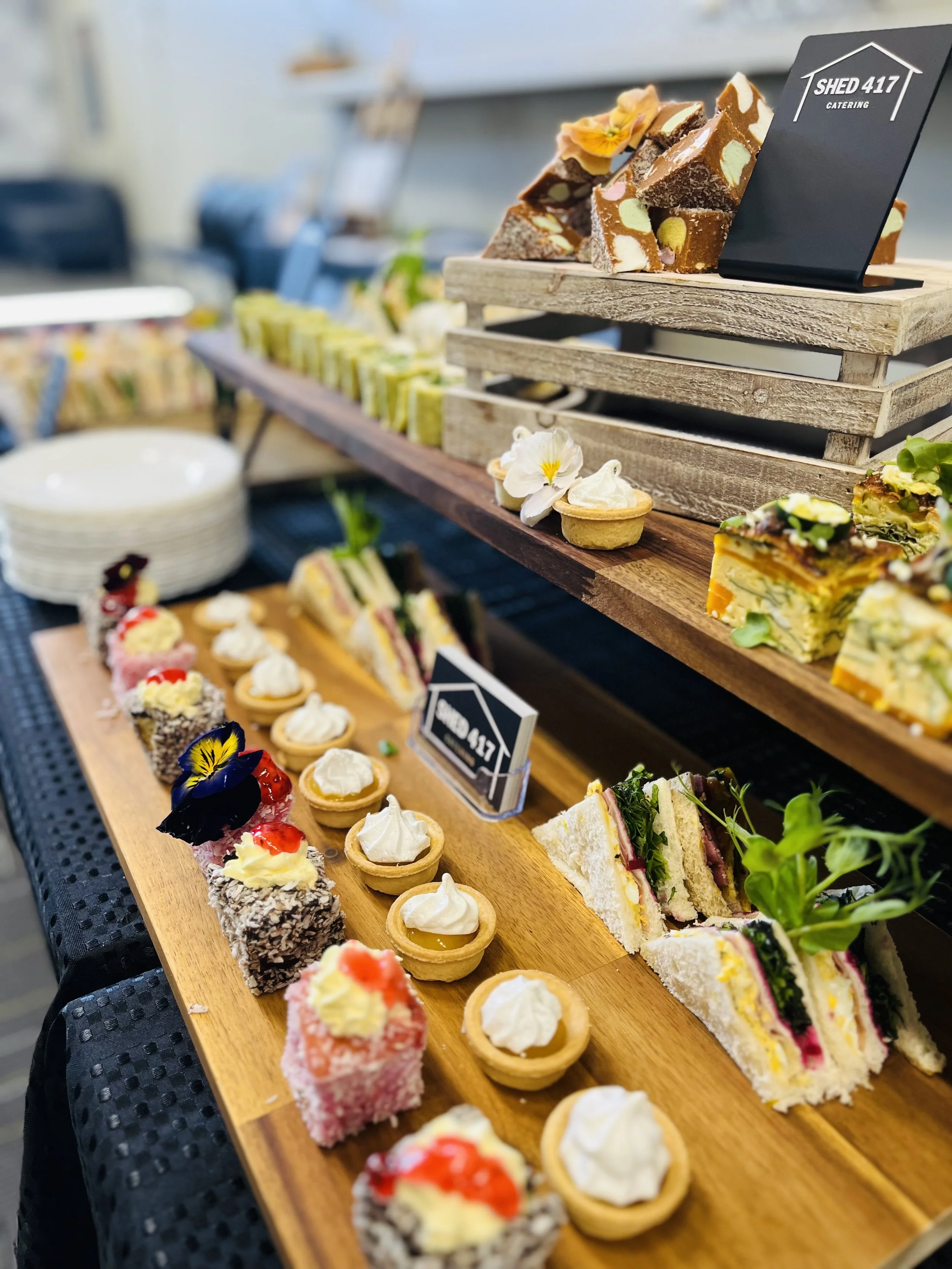 A wooden serving board displaying assorted desserts including small cakes, tartlets with whipped cream, and colorful layered sandwiches at a catered event.