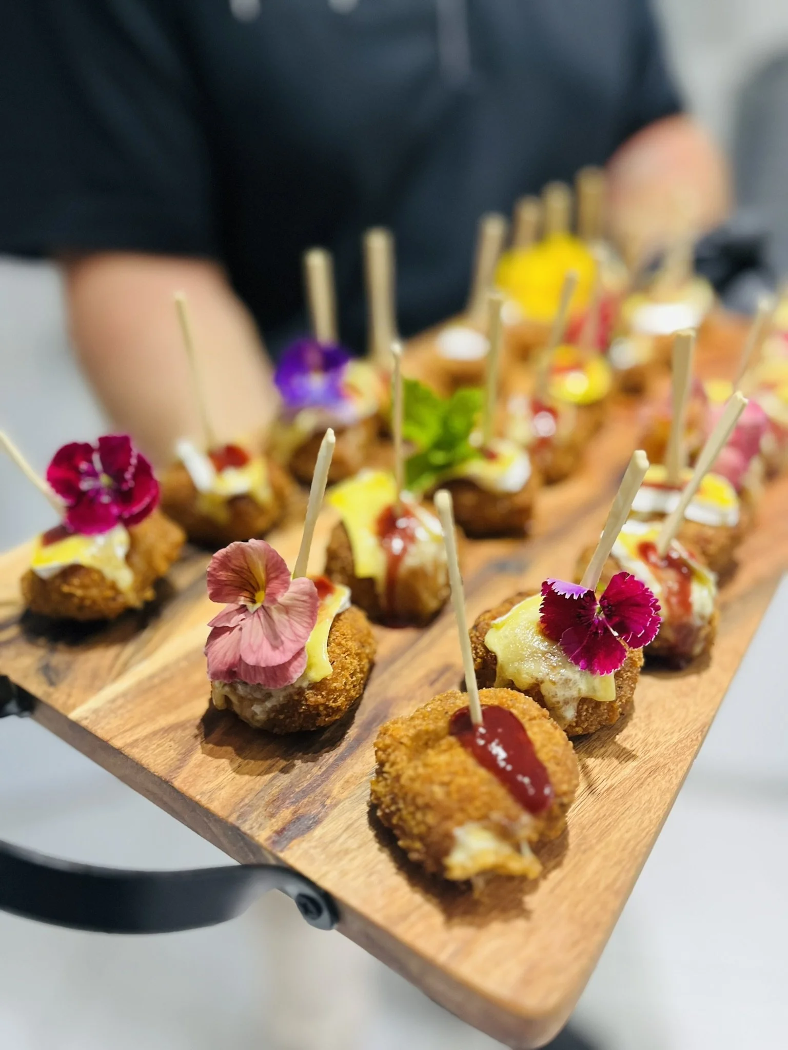 A person holding a wooden serving tray with several pieces of fried food topped with cheese, sauce, and edible flowers.