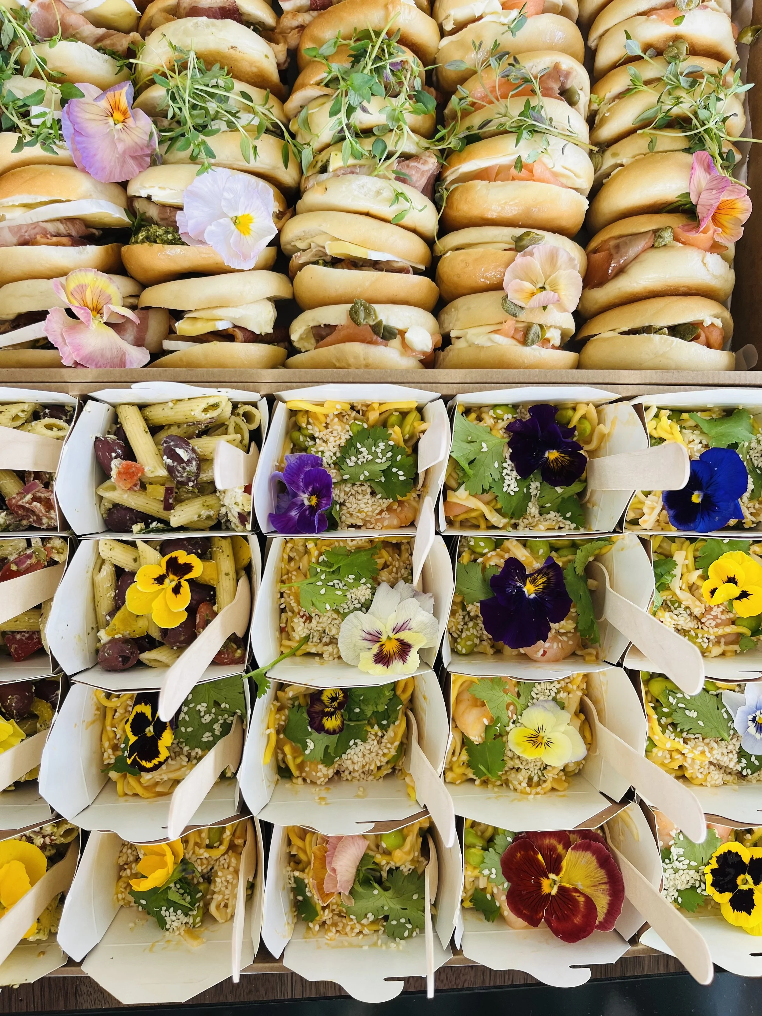 Tray of assorted mini sandwiches with flowers and herbs, and small boxes of pasta salads decorated with edible flowers.