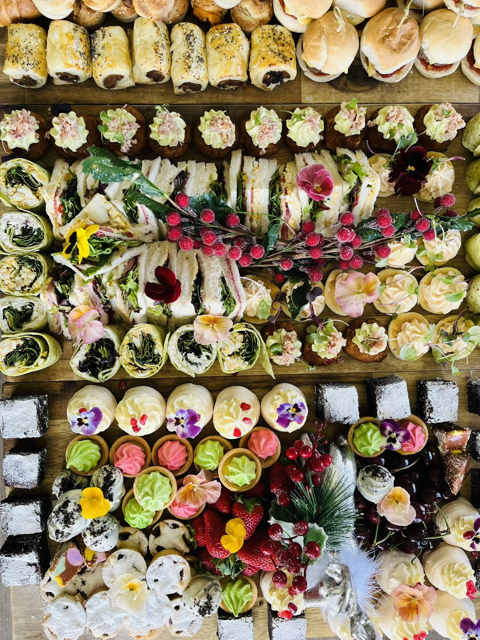 Assorted Christmas-themed desserts including mini sandwiches, cupcakes, cookies, and petit fours, decorated with edible flowers, holly, berries, and festive decorations, arranged on a wooden surface.