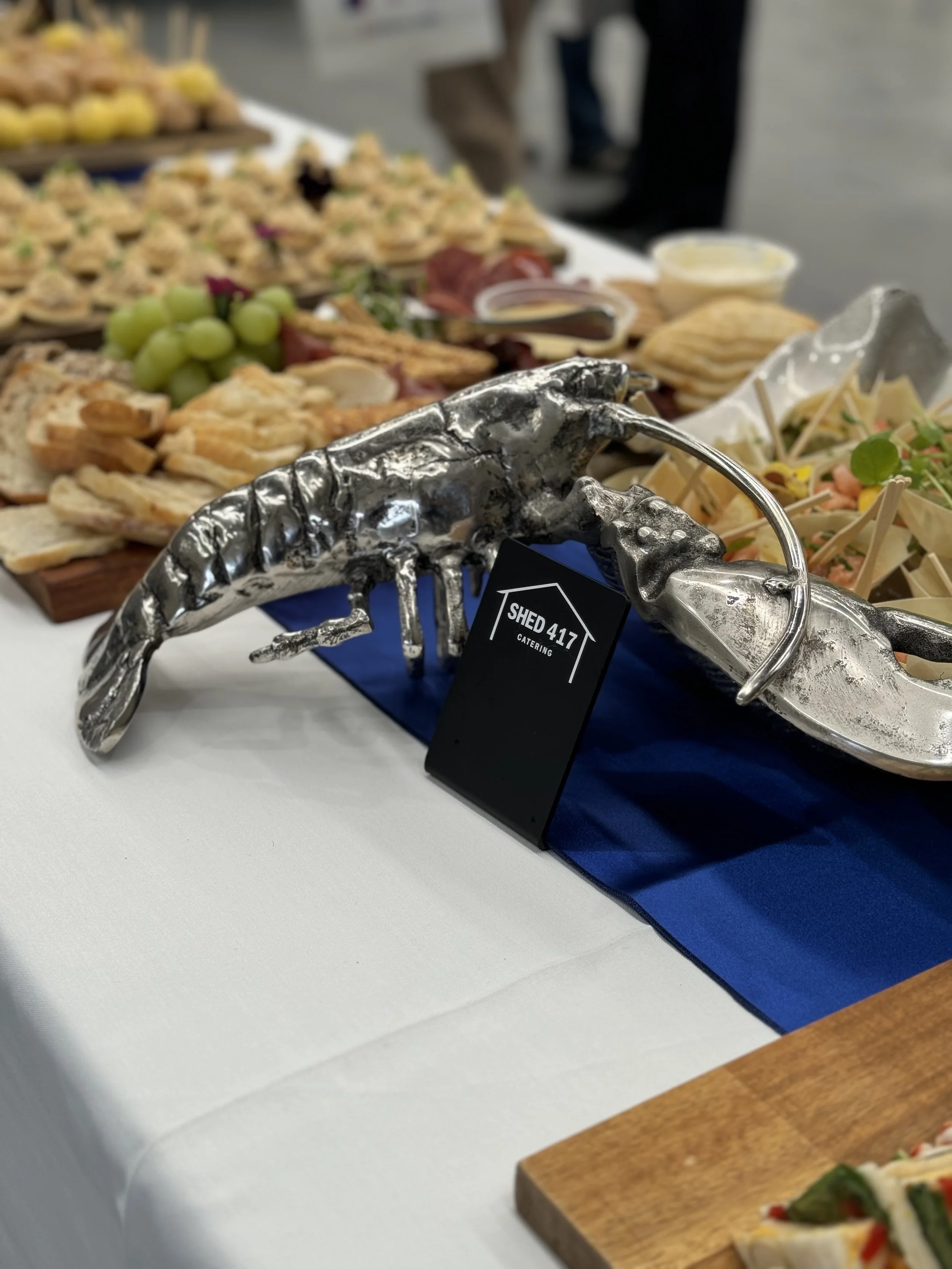 A catering display with a silver lobster sculpture holding a black card with the text 'SHED 417 CATERING', surrounded by various appetizers including grapes, sliced bread, and small bowls of dips, on a table with a white tablecloth and a blue runner.