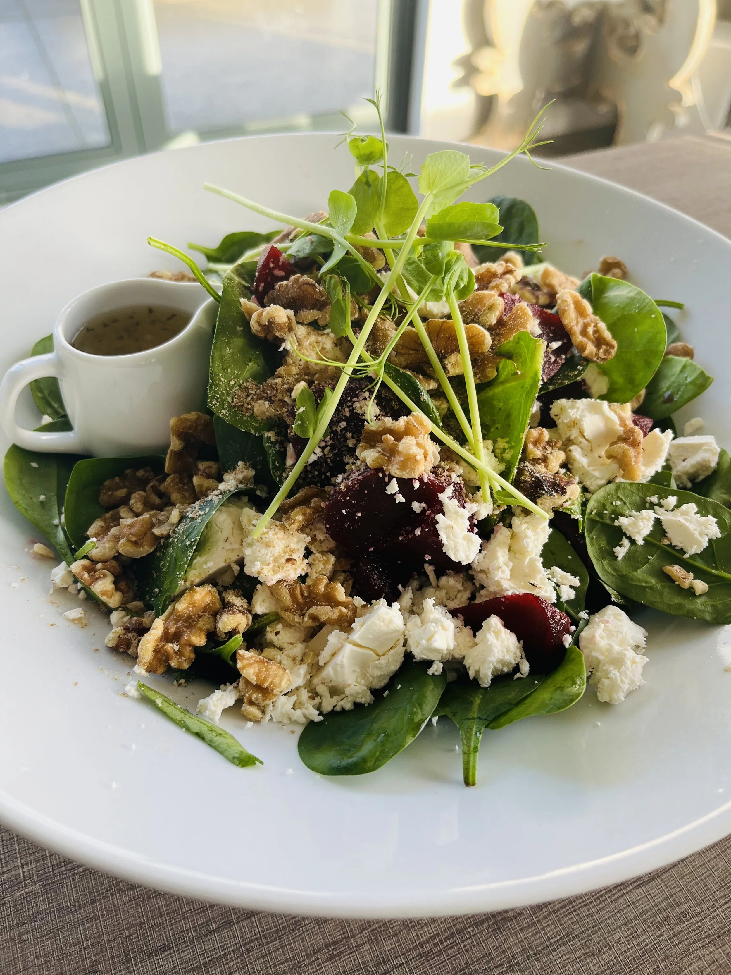 Mixed salad with greens, crumbled cheese, walnuts, cherry beets, microgreens, and a small cup of dressing on a white plate.