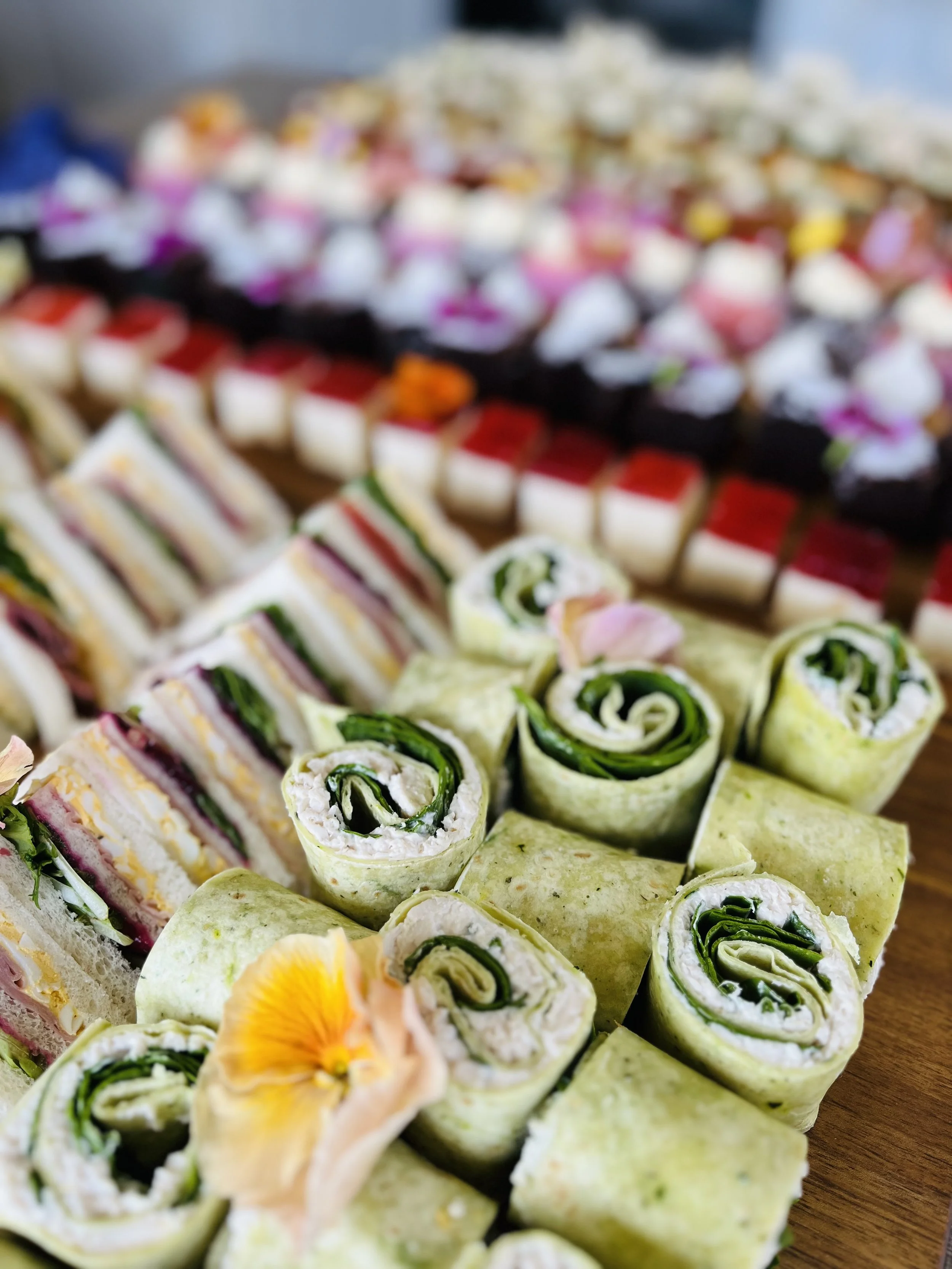 Tray of assorted pinwheel sandwiches and small pastry squares, with a blurred background of more desserts or appetizers.