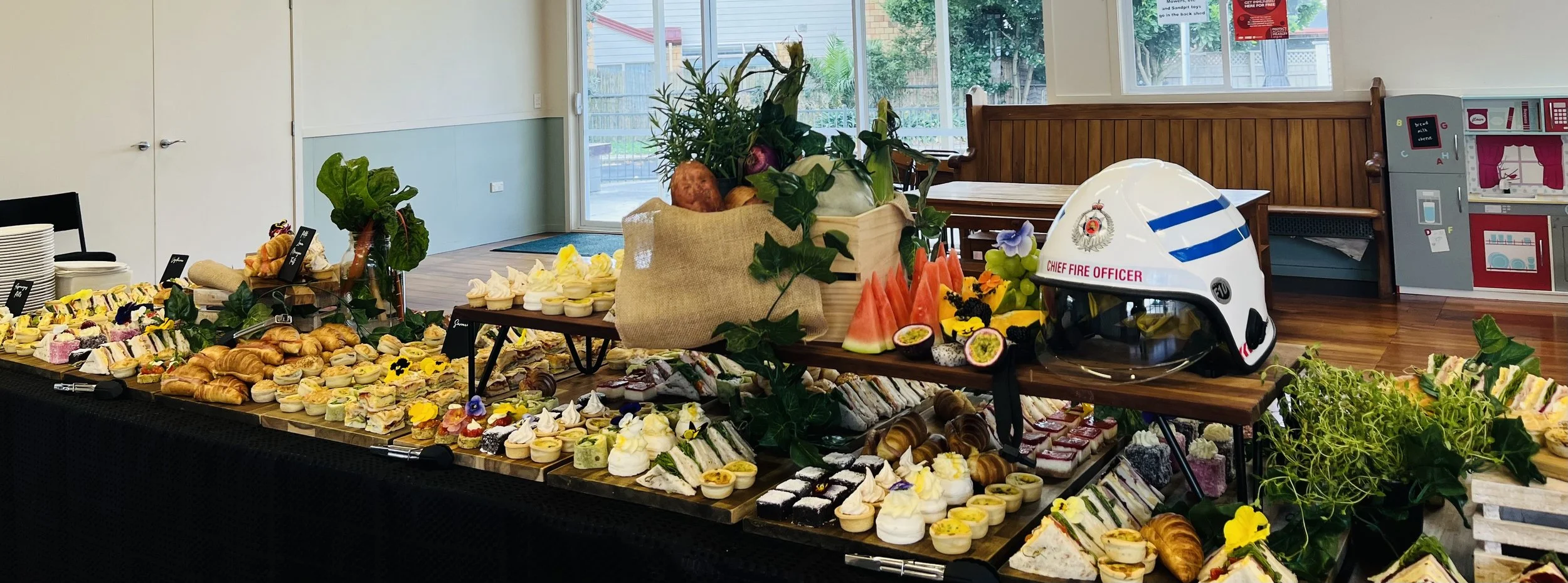 A buffet table with sandwiches, pastries, and fruit, decorated with greenery and flowers. A firefighter helmet is on the table. In the background, there is a wooden bench and children's play kitchen in a room with large windows.
