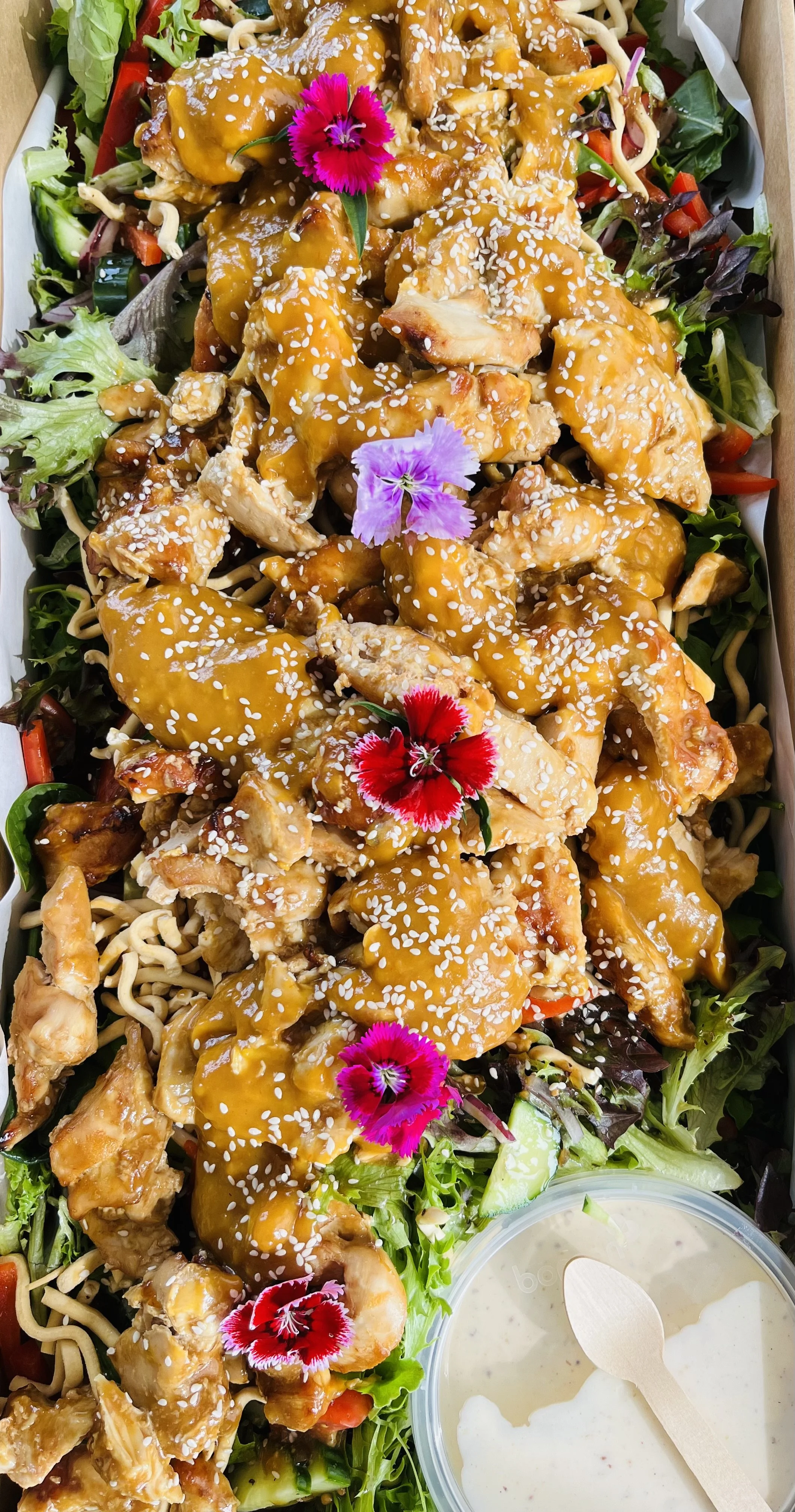 Salad topped with diced cooked chicken pieces covered in orange sauce, sprinkled with sesame seeds, garnished with edible flowers, with a side of creamy dressing.