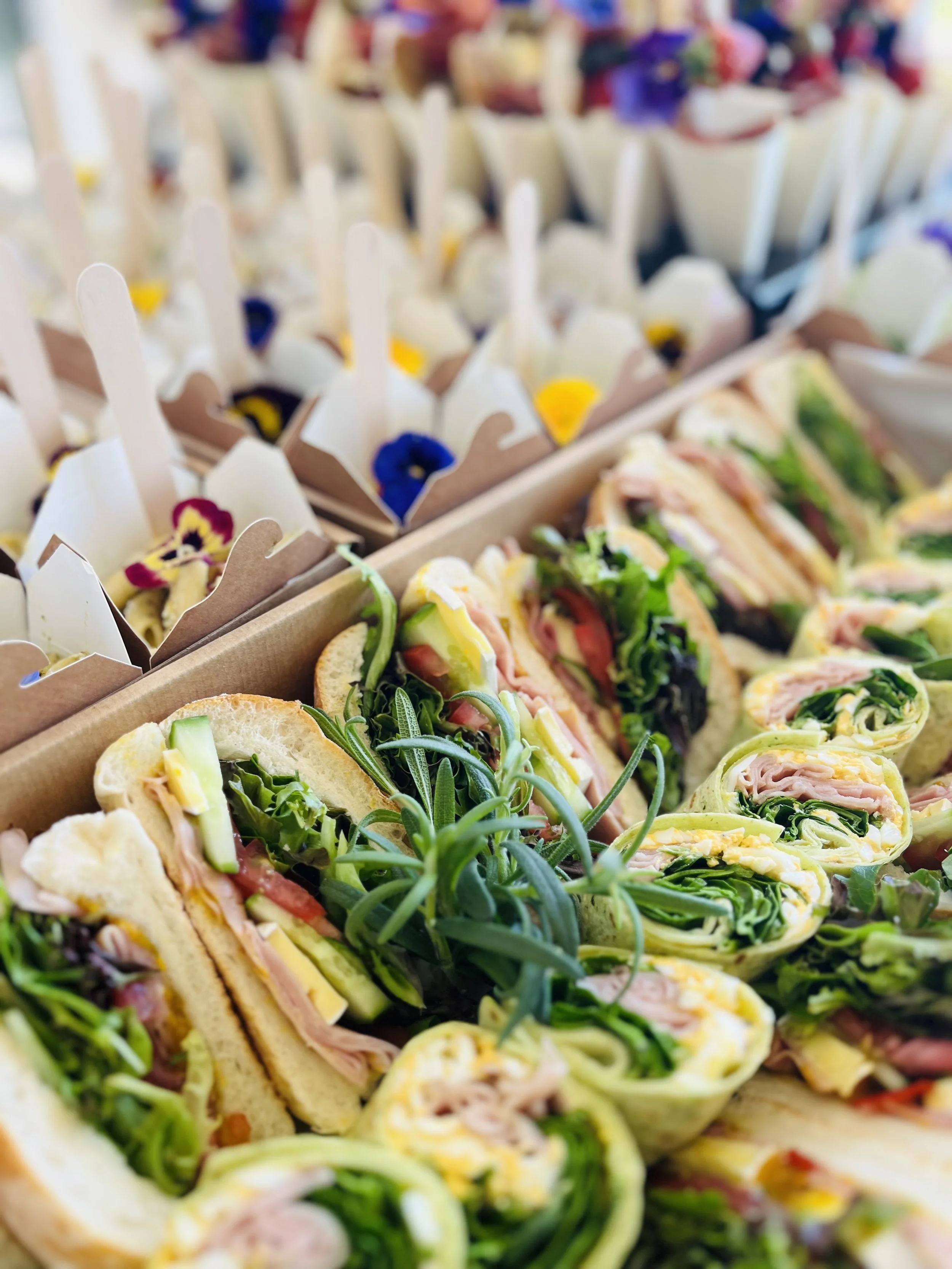Assorted sandwiches and appetizers on a tray, including veggie wraps and small cups with spoons, decorated with edible flowers.