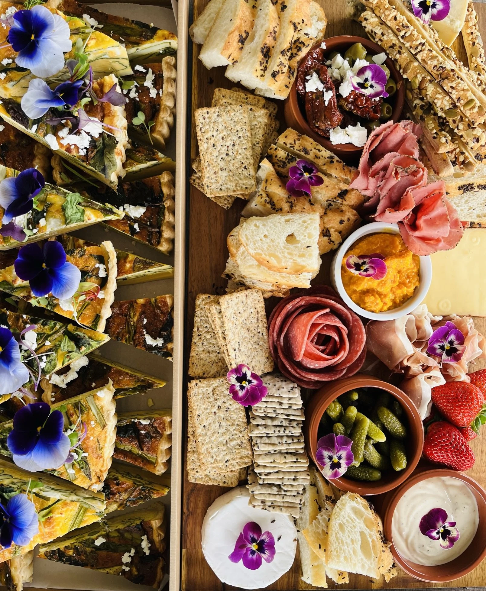 A variety of colorful appetizers including vegetable tart slices garnished with edible flowers on the left, and on the right, assorted cheeses, cured meats, crackers, olives, strawberries, pickles, and dipping sauces, all decorated with edible flower