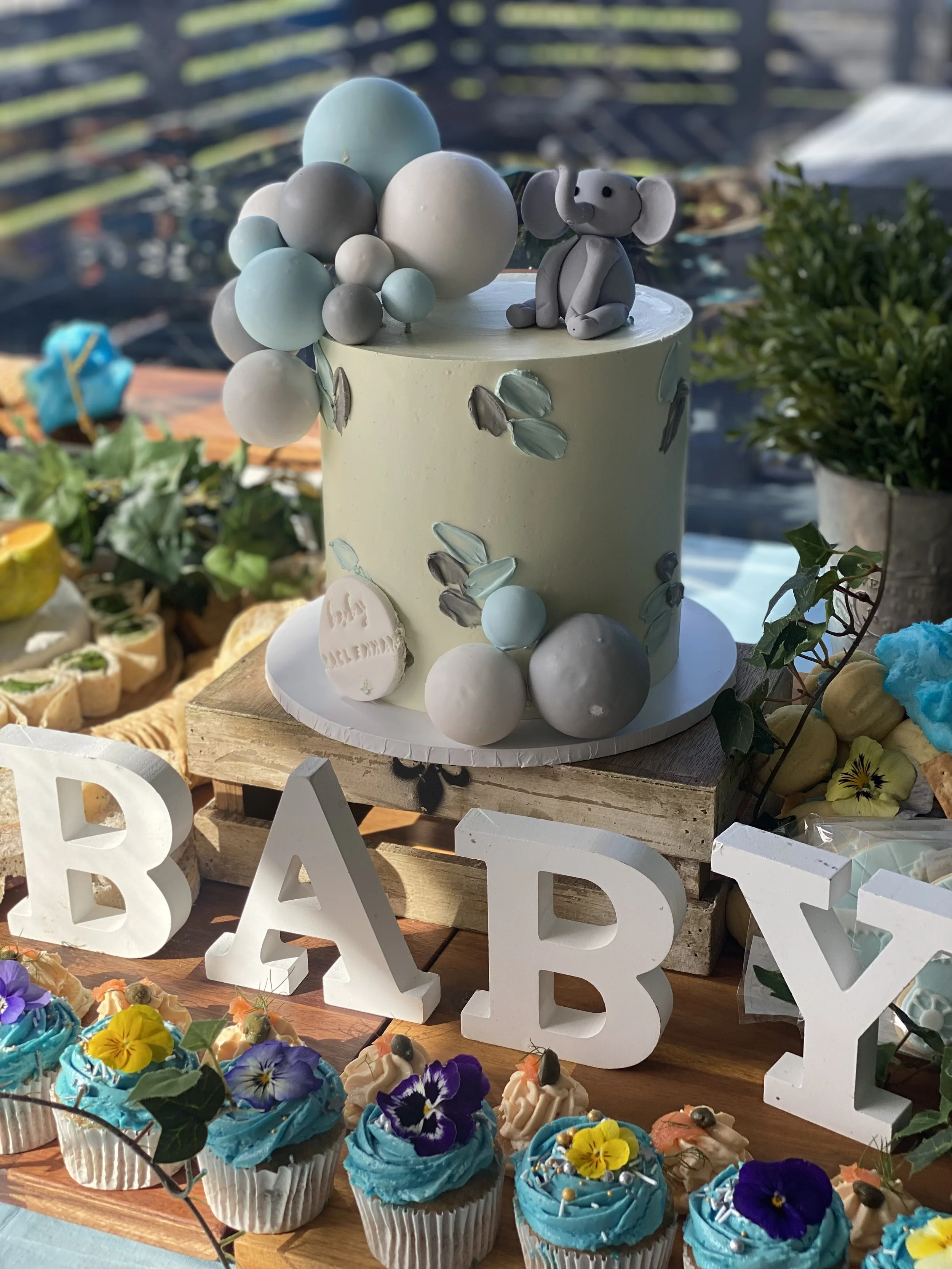 Baby-themed birthday cake with pastel decor, including a small elephant figurine, balloons, and cupcakes decorated with flowers and edible toppers, spelling out 'BABY'.