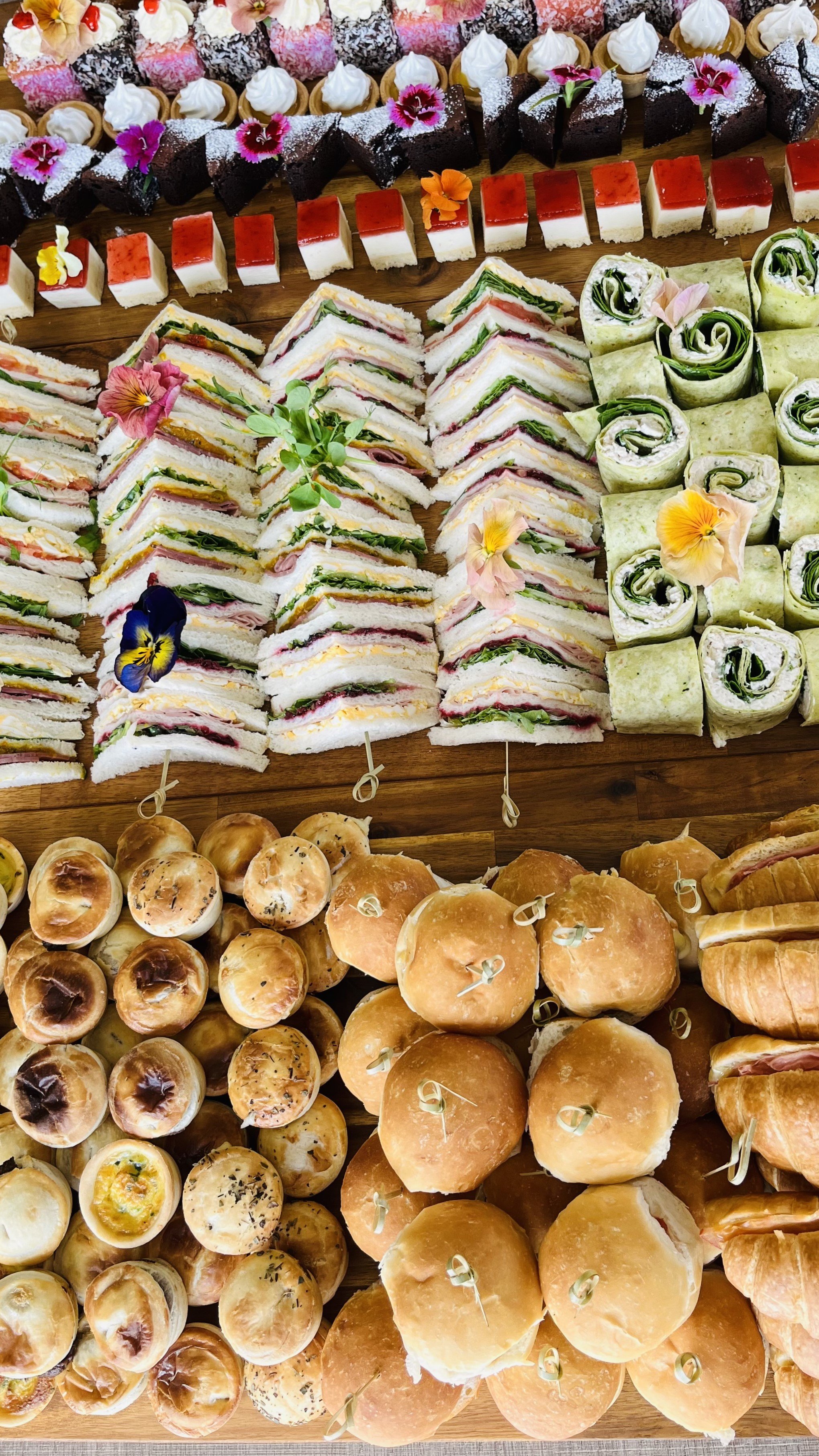 Assorted finger foods including mini sandwiches, wraps, sliders, scones, and various desserts arranged on a wooden table with edible flowers for decoration.